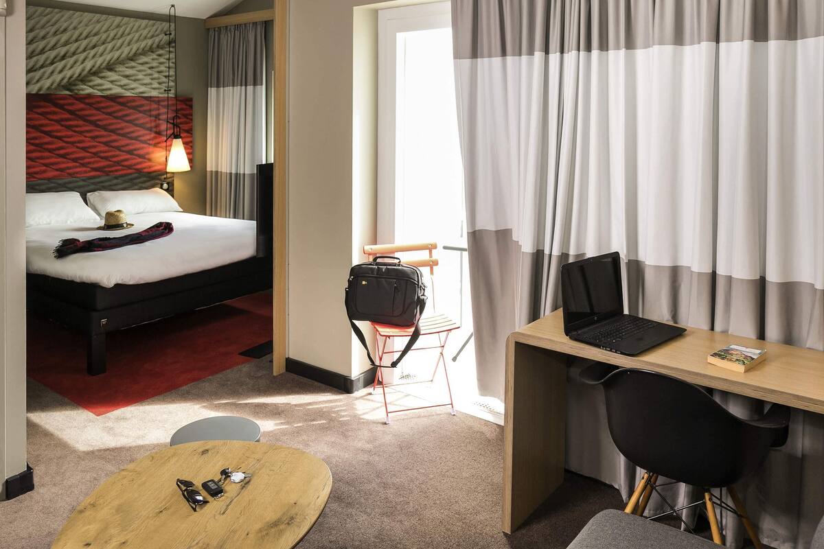 Une chambre d’hôtel avec un lit, un bureau avec un ordinateur portable, une chaise, une petite table avec des lunettes de soleil et un porte-clés, et une valise posée sur une chaise.
