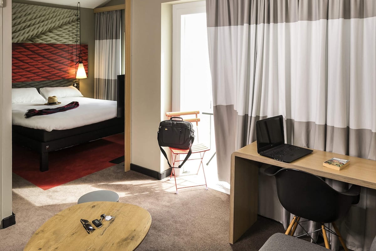 Habitación de hotel con cama, escritorio con una computadora portátil, una silla, una mesita con gafas de sol y un llavero, y una maleta sobre una silla.