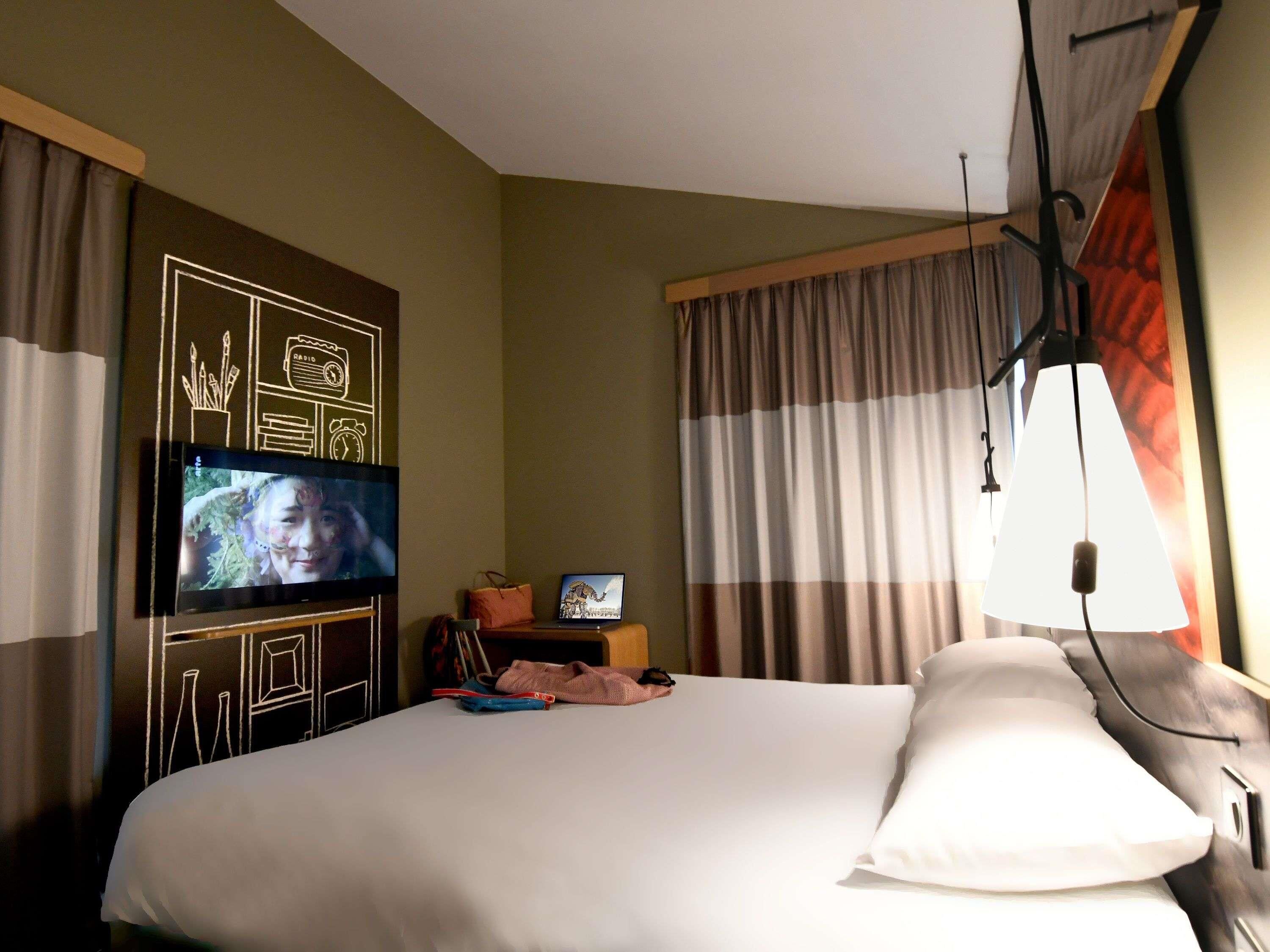 Une chambre d’hôtel avec un lit, une télévision, un bureau avec un ordinateur portable et des rideaux.