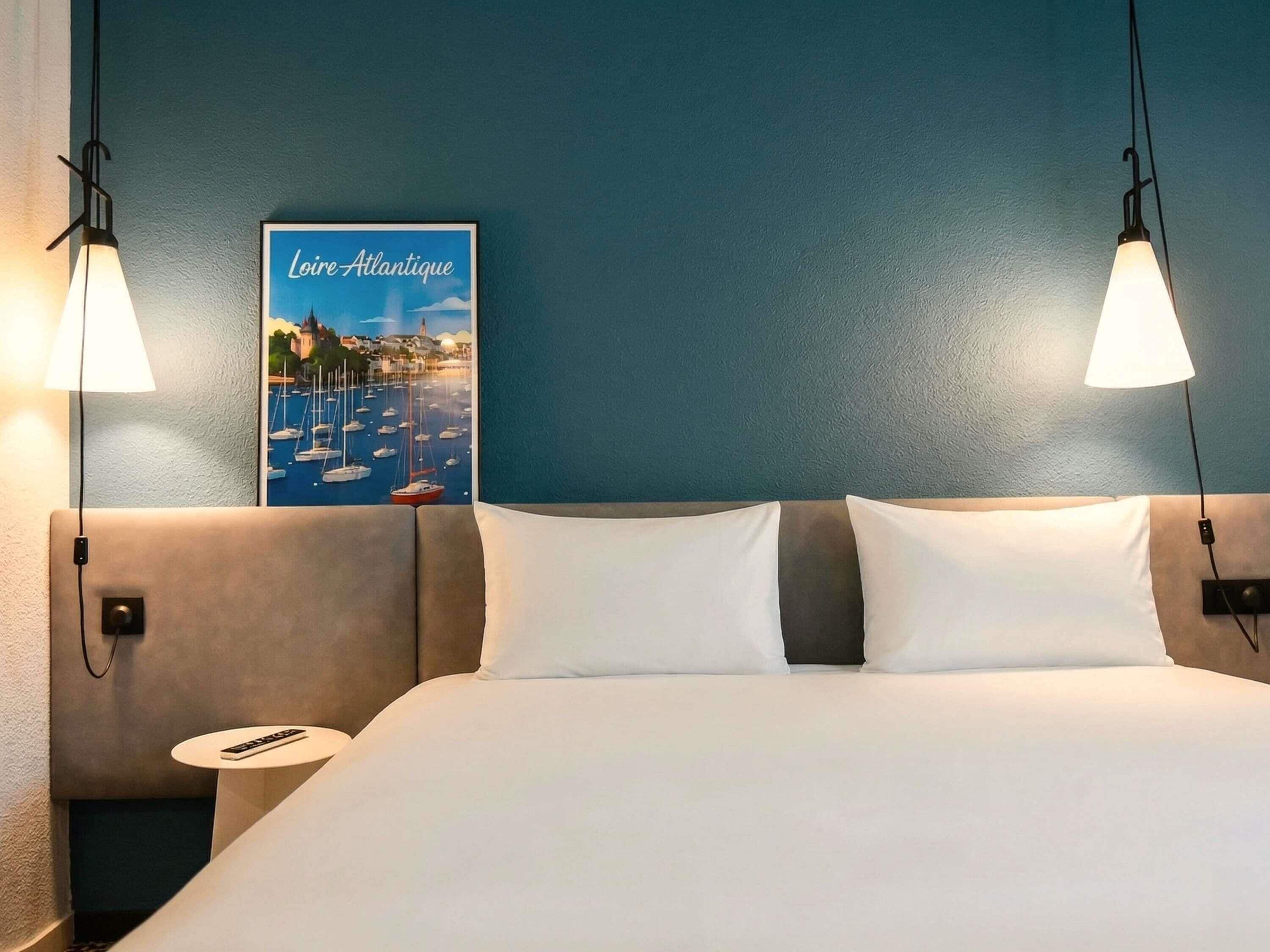 A hotel room with a bed, two bedside lamps, a nightstand, a remote control, and a poster of a coastal scene.