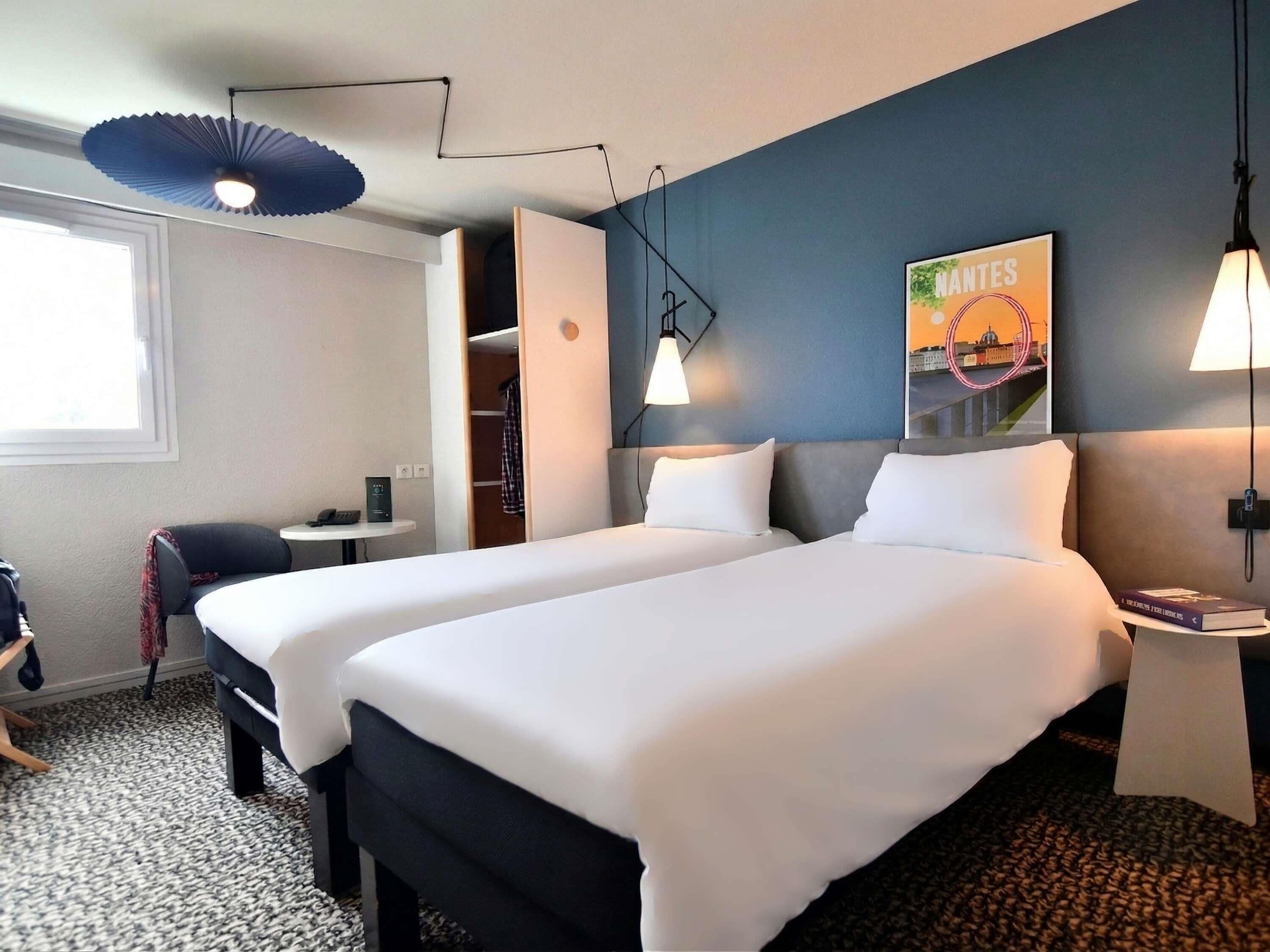 Habitación de hotel con dos camas, un escritorio, una silla, una mesita, una lámpara y un estante en la pared.