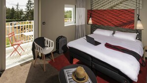 Ein Hotelzimmer mit Bett, Schreibtisch, Stuhl, Koffer und einem Balkon mit Blick auf Bäume und Autos.