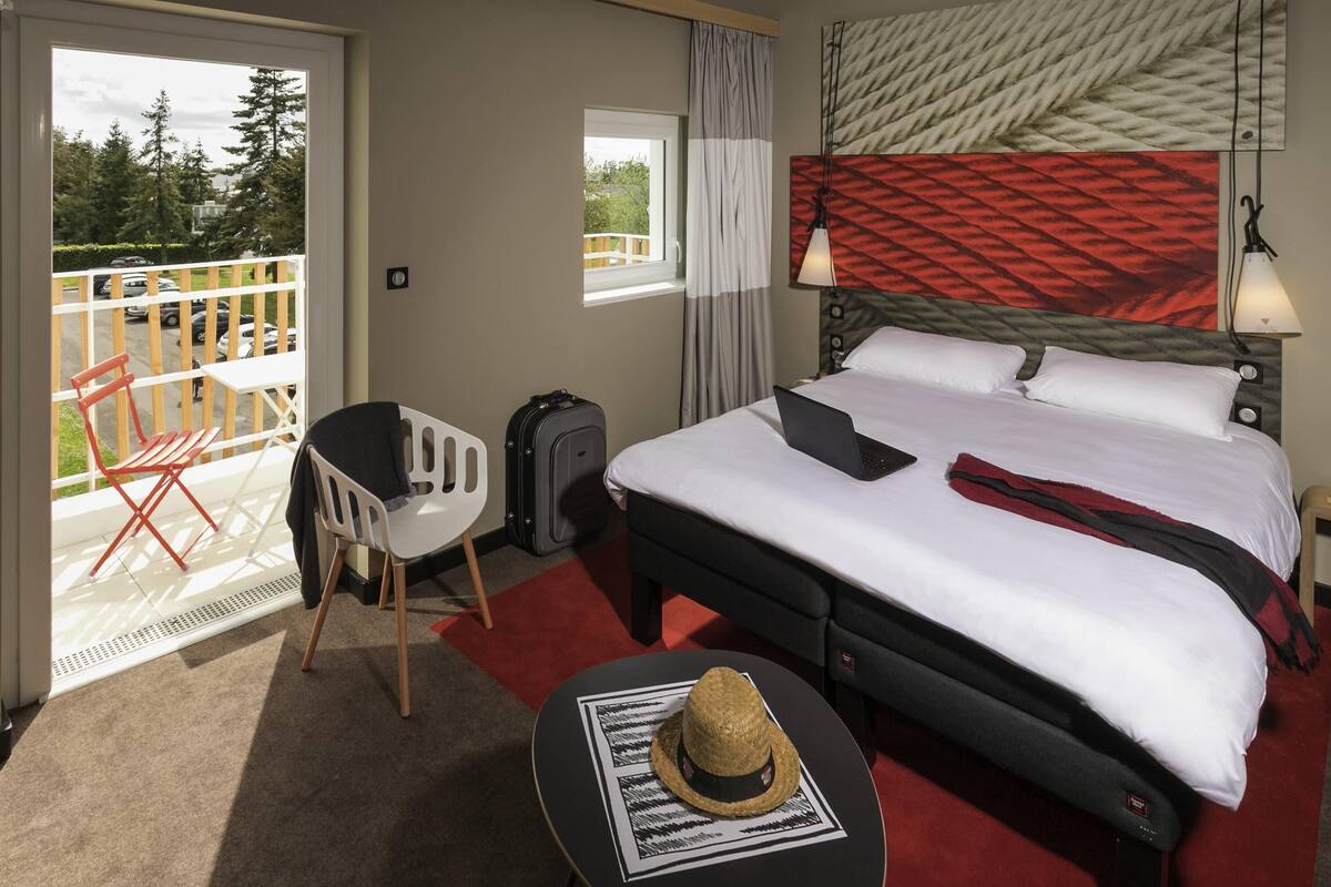 Une chambre d’hôtel avec un lit, un bureau, une chaise, une valise et un balcon donnant sur des arbres et des voitures.