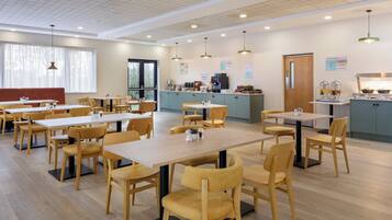 Un grand espace à manger avec plusieurs tables et chaises, un coin cuisine doté d’un comptoir et une grande fenêtre agrémentée de rideaux.