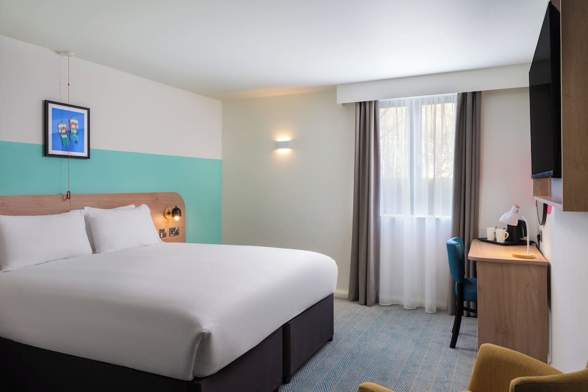 A hotel room with a large bed, a desk with a lamp, a chair, and a TV on the wall.