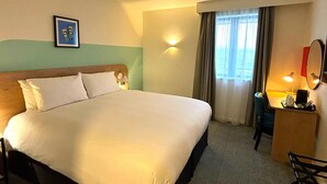 Una habitación de hotel con una cama grande, un escritorio con silla, una lámpara y un cuadro en la pared.