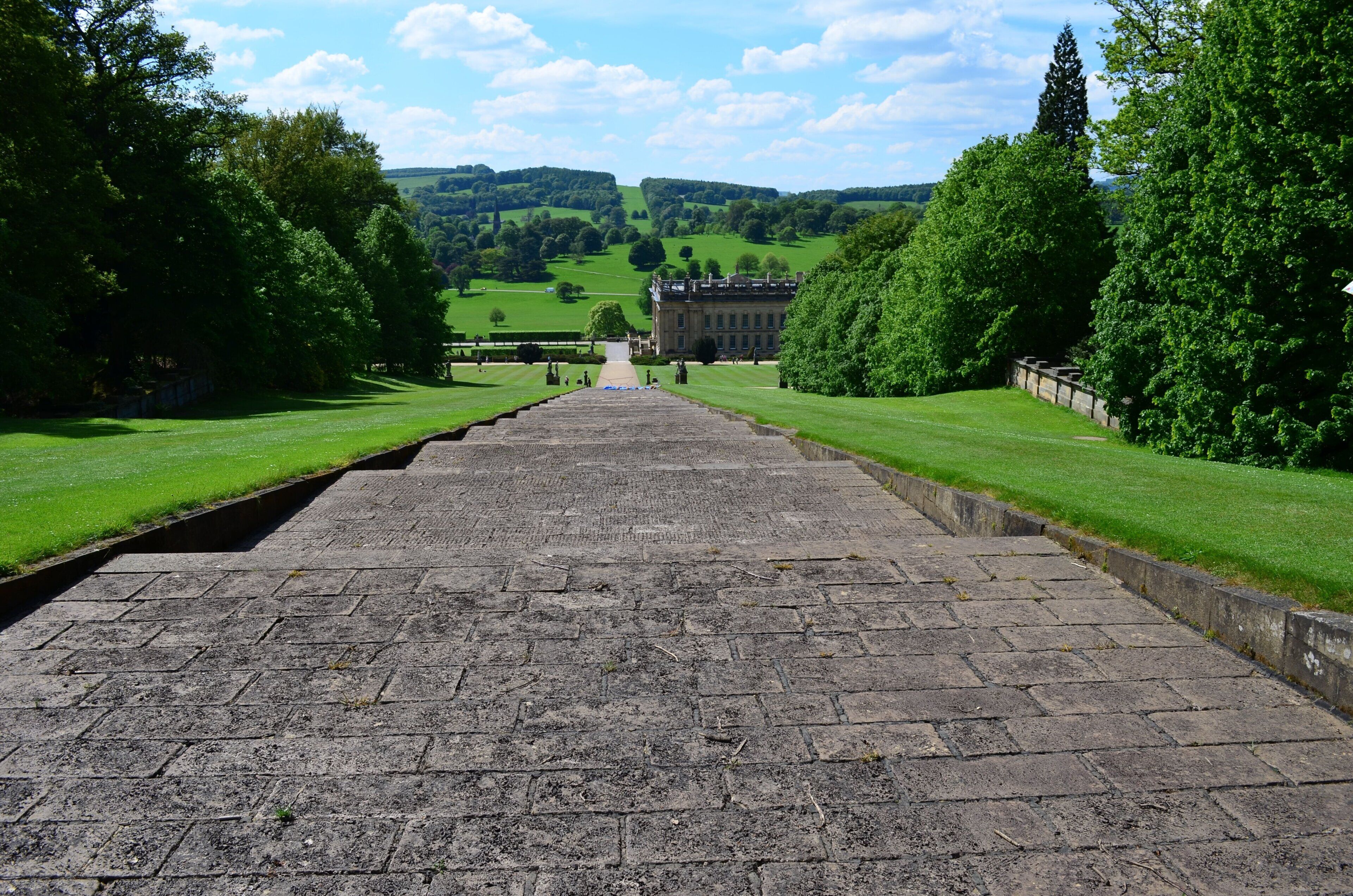 A paved pathway leading to a large building with a formal entrance, surrounded by well-maintained lawns and trees.
