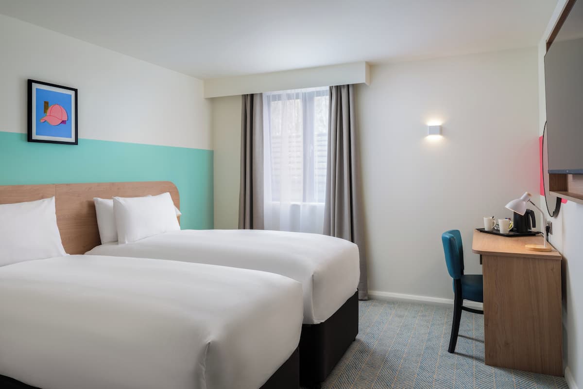 A hotel room with two beds, a desk with a chair, a picture on the wall, and a window with curtains.