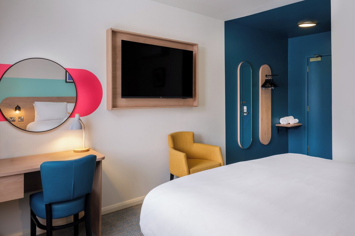 A hotel room with a bed, a desk with a chair, a TV, a mirror, and a blue wall with a door.