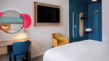 A hotel room with a bed, a desk with a chair, a TV, a mirror, and a blue wall with a door.