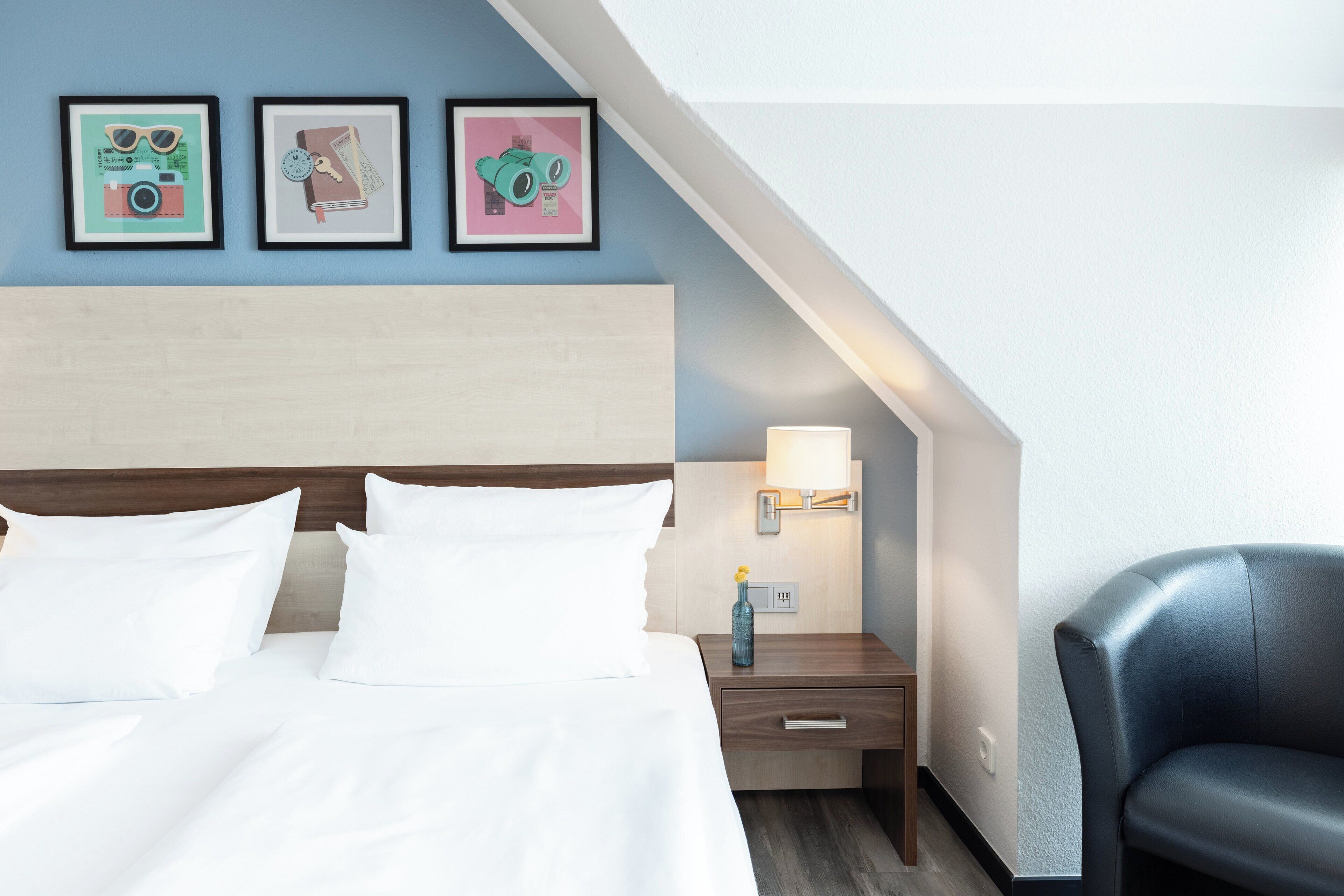 Habitación de hotel con cama, mesita de noche, lámpara, silla y tres cuadros en la pared.