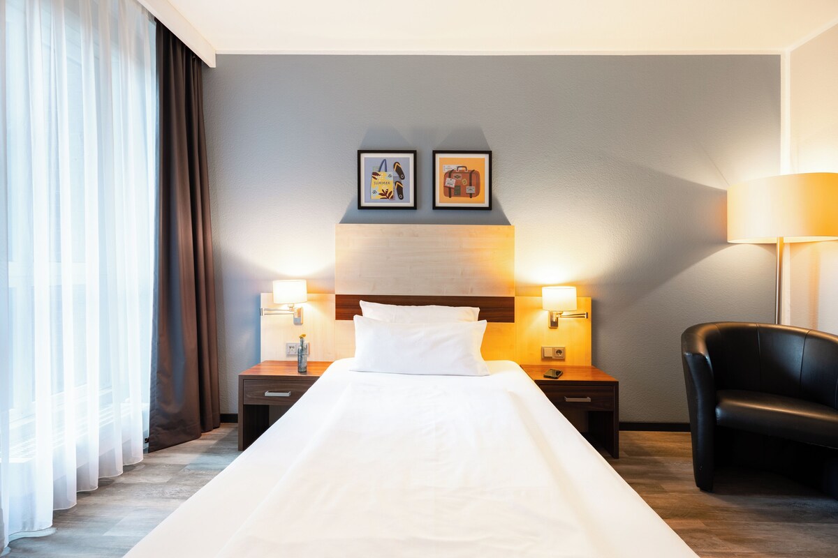 Une chambre d’hôtel avec un lit, deux tables de chevet avec des lampes, une chaise et deux tableaux encadrés au mur.