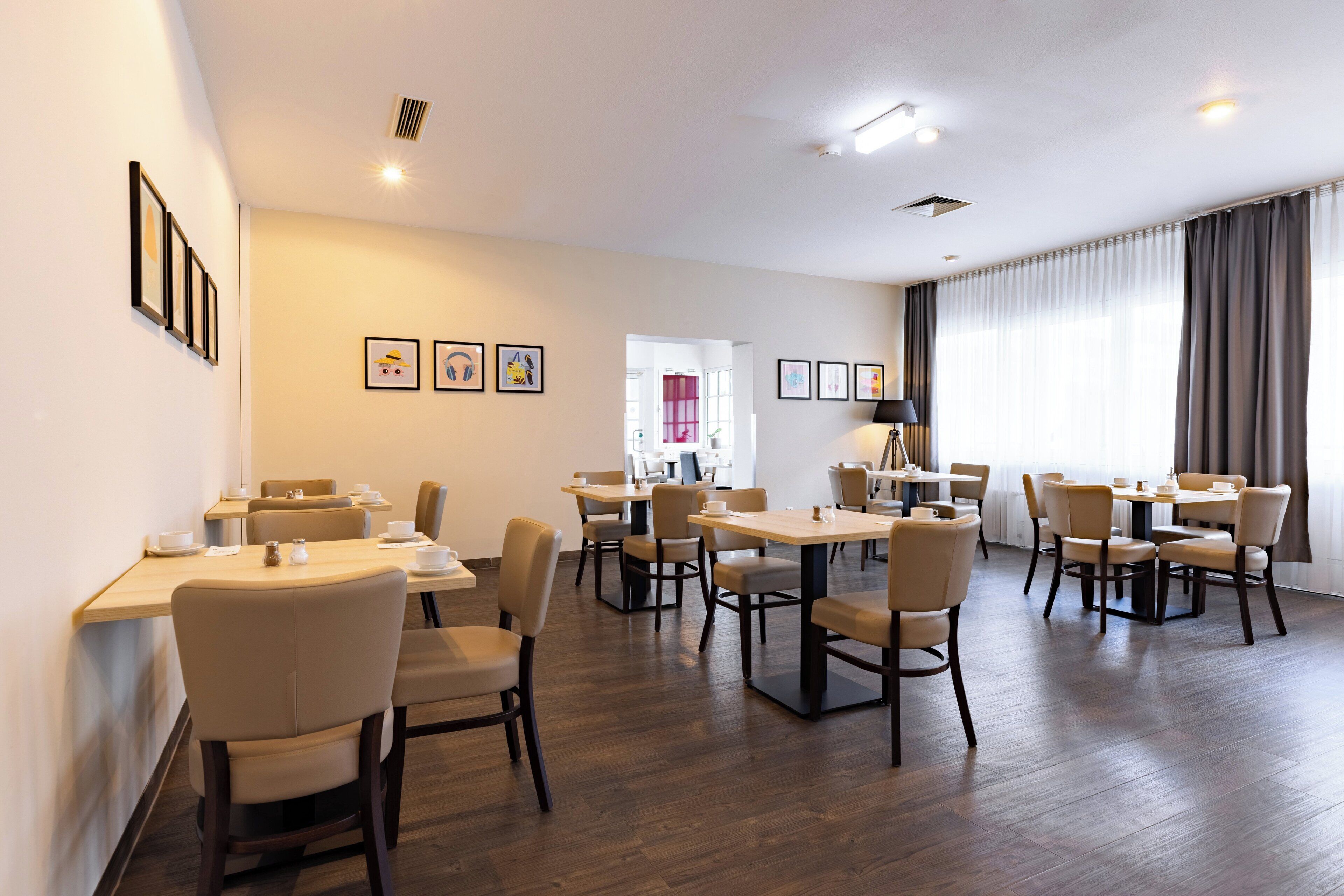 Un espace repas moderne, doté de plusieurs tables et chaises, avec des œuvres d’art encadrées accrochées aux murs et offrant une vue sur l’extérieur grâce à de grandes fenêtres.