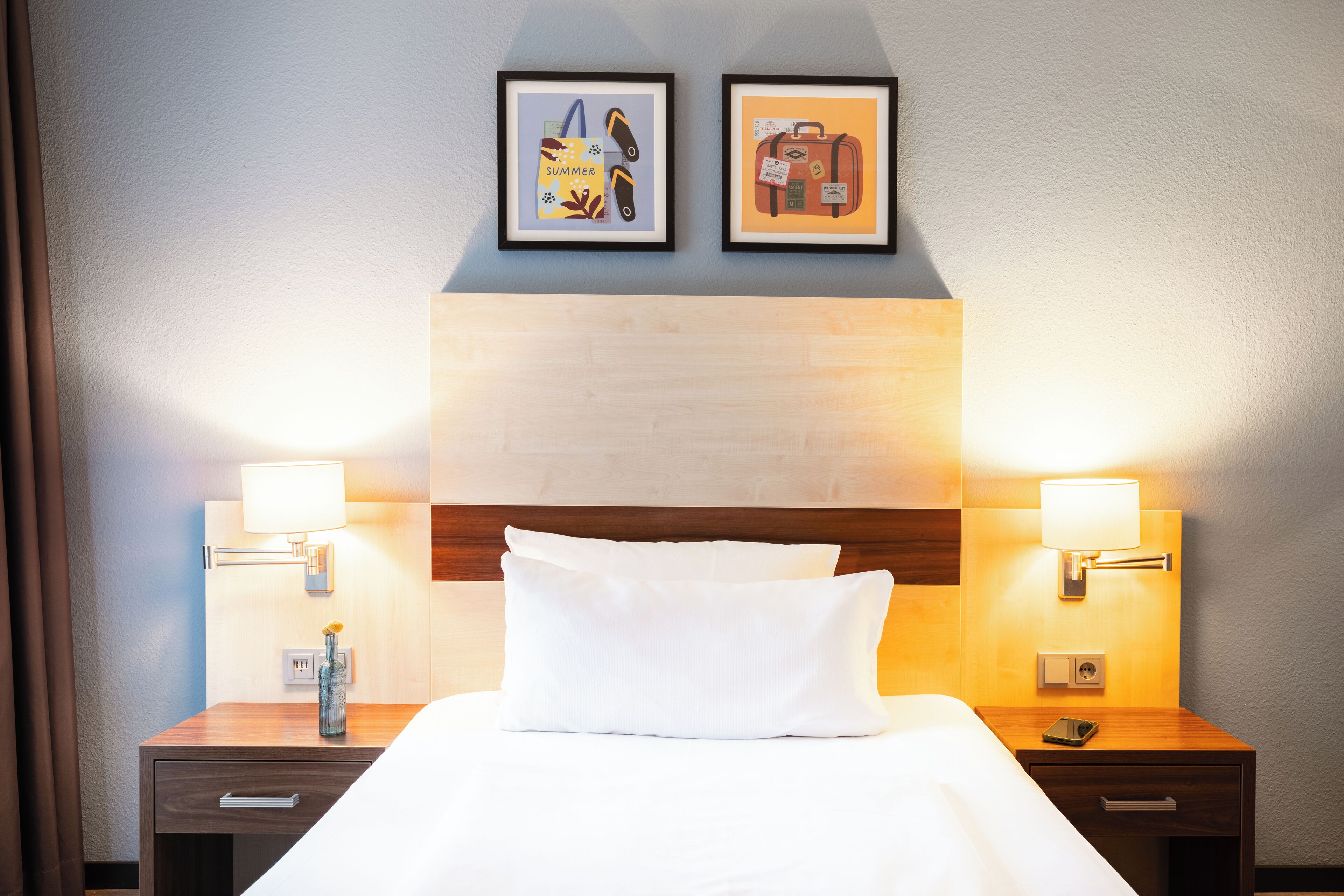 Ein Hotelzimmer mit einem ordentlich bezogenen Bett, zwei Nachttischen mit Lampen und zwei gerahmten Bildern an der Wand.
