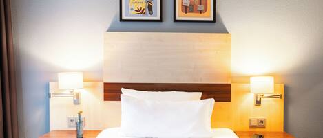 Ein Hotelzimmer mit einem ordentlich bezogenen Bett, zwei Nachttischen mit Lampen und zwei gerahmten Bildern an der Wand.
