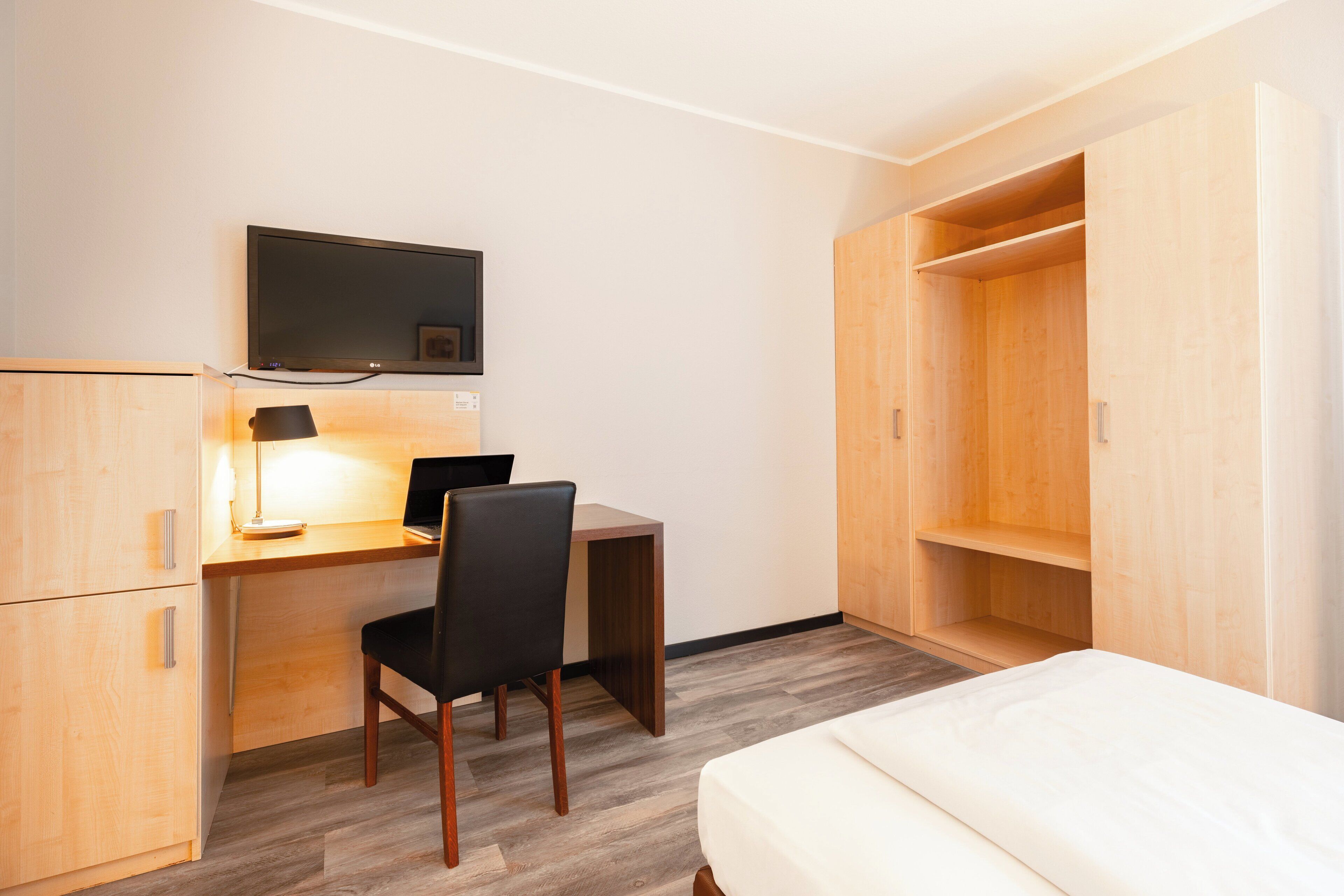 Une chambre d’hôtel compacte comprenant un bureau en bois, une chaise, un lit, un téléviseur fixé au mur et une armoire.