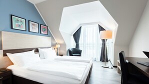 A modern hotel room with a large bed, a desk, and a chair. There are framed pictures on the wall and a lamp on the desk.