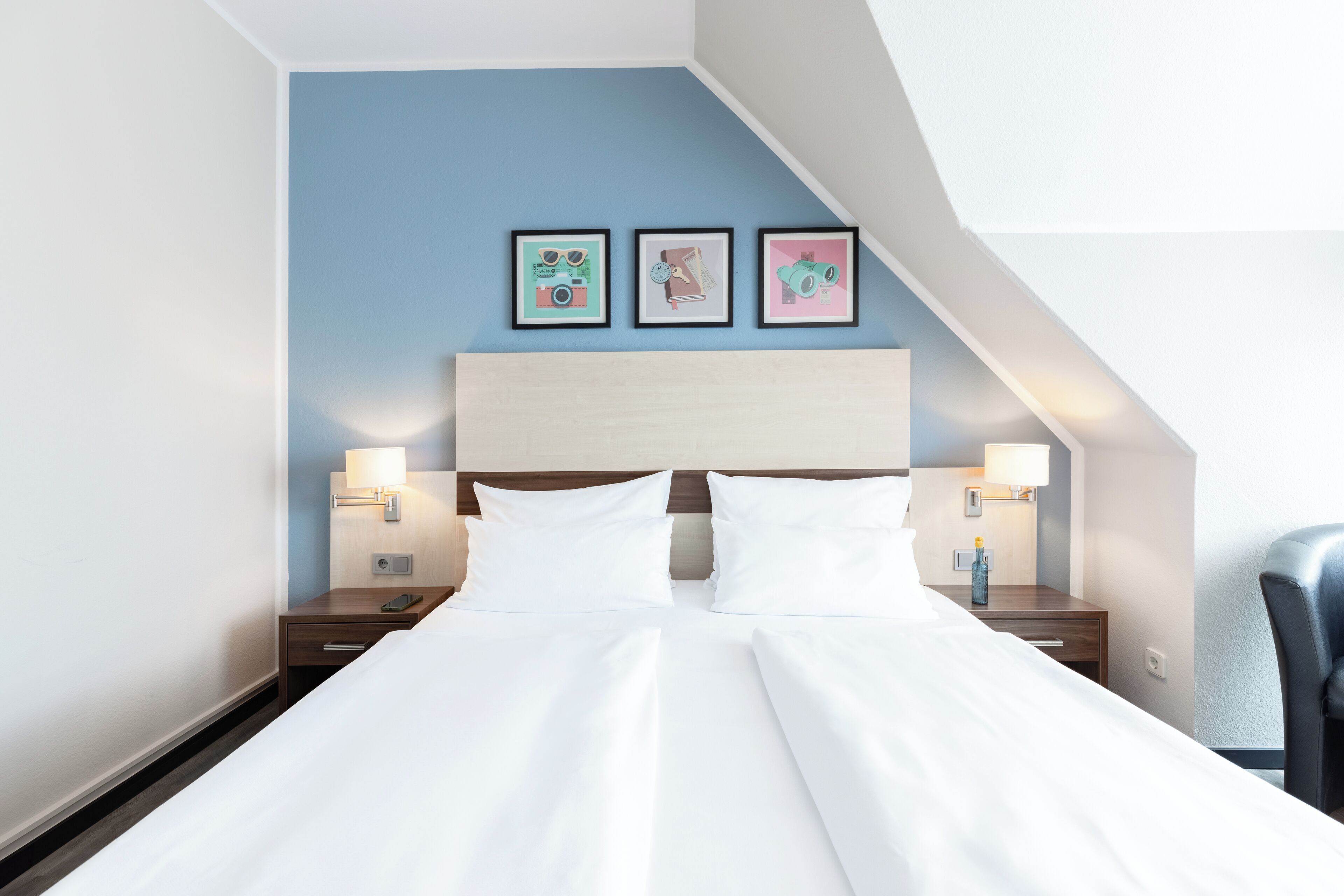 A hotel room with a large bed, two bedside tables with lamps, and a headboard. There are three framed pictures on the wall above the bed.