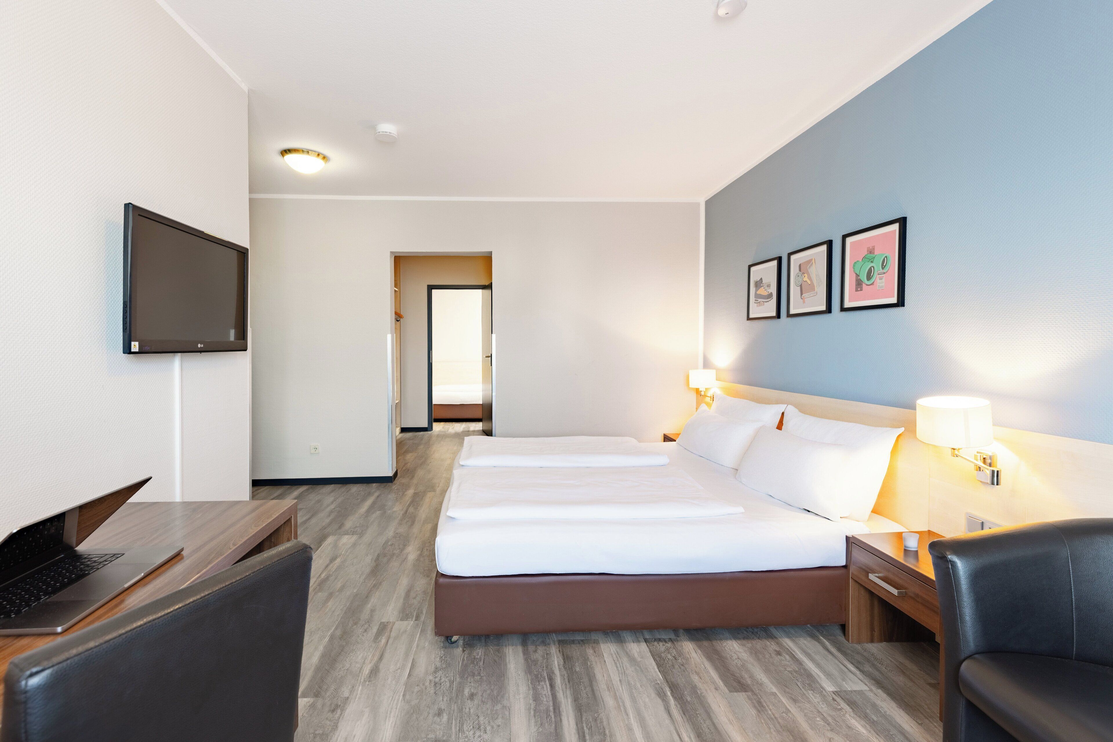A hotel room with a bed, a desk with a laptop, a TV, and a nightstand with a lamp.