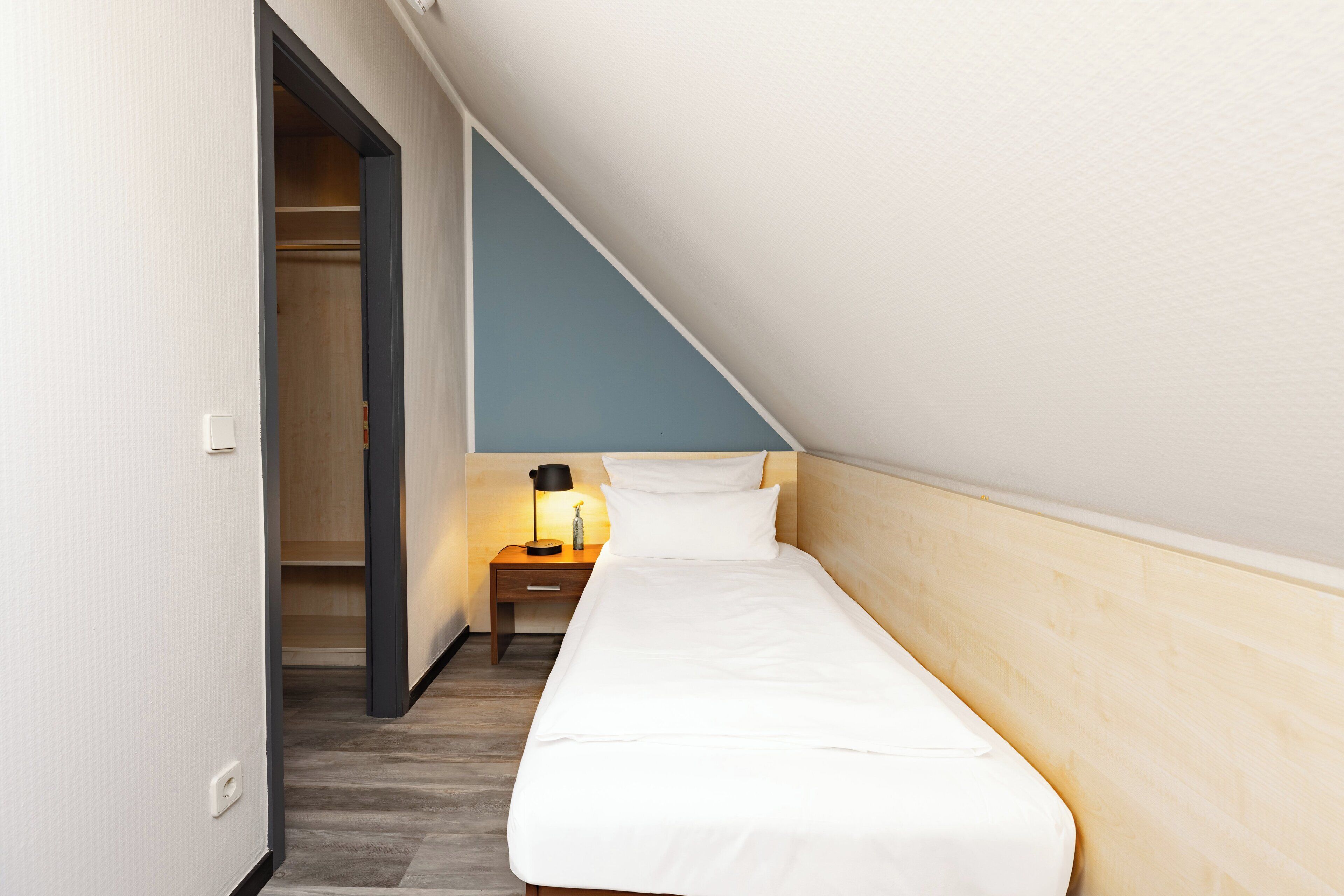 A small, neatly arranged bedroom with a single bed, a bedside table with a lamp, and a wooden wardrobe.