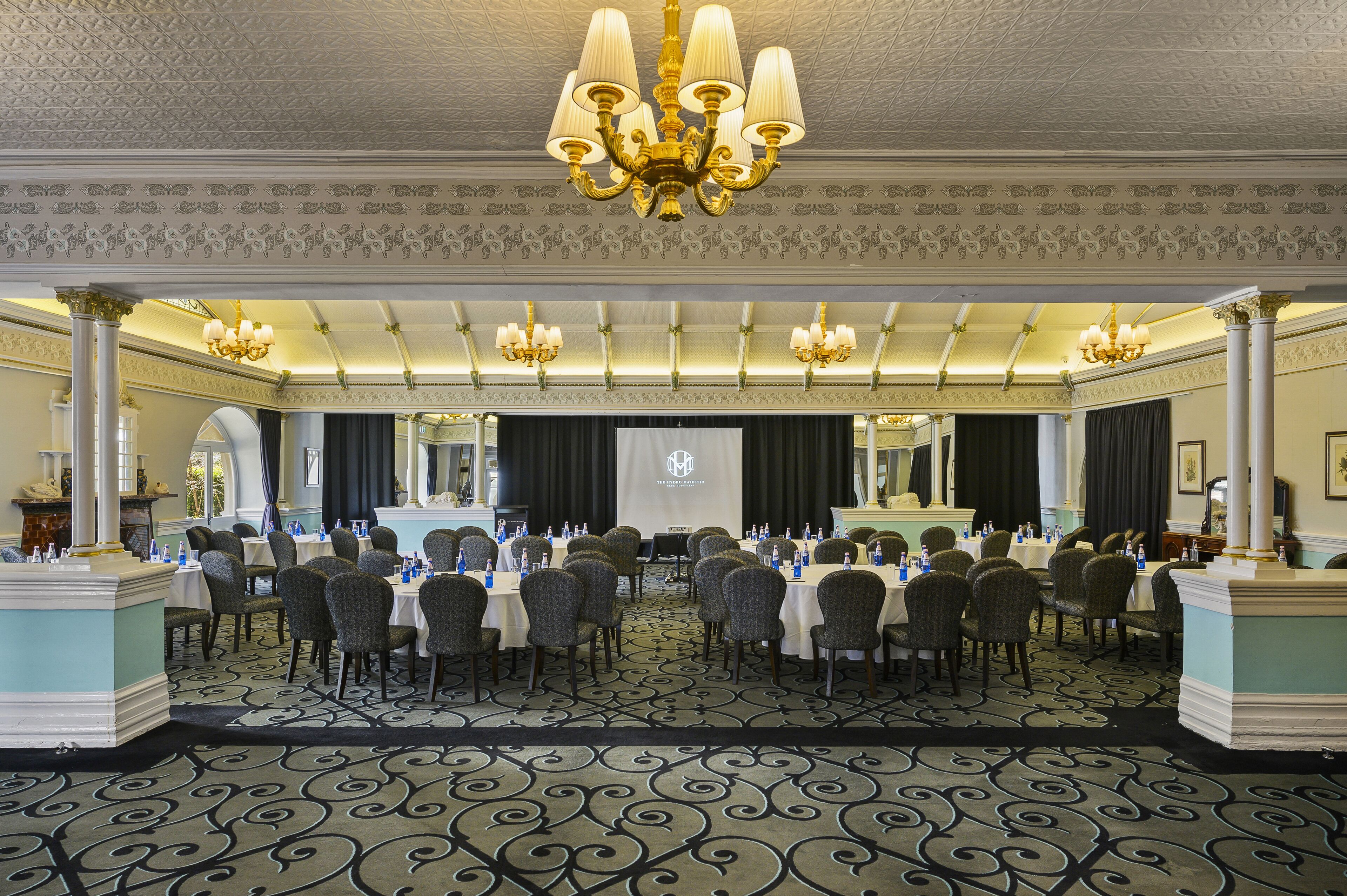 A banquet hall with round tables, chairs, chandeliers, and a projector screen.