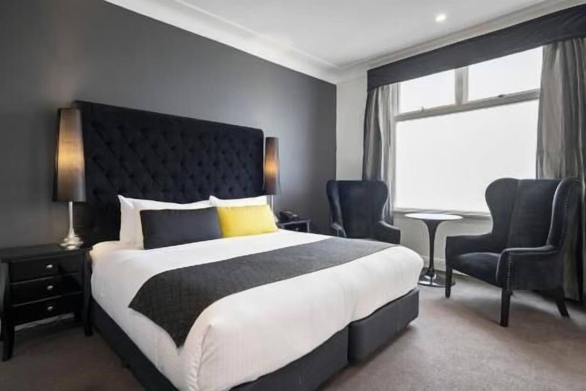 A hotel room with a large bed, a bedside table, a chair, a small round table, and a window with curtains.