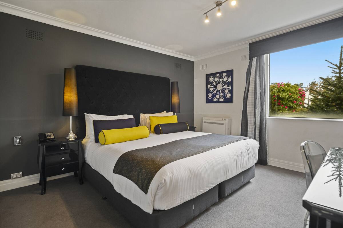 A modern bedroom with a large bed, bedside tables, a desk, and a window with a view of greenery.
