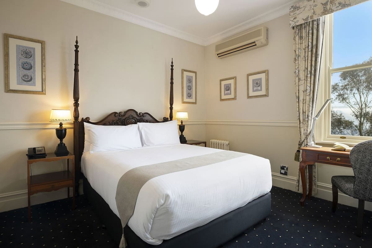A hotel room with a four-poster bed, two bedside tables with lamps, a desk with a chair, and framed pictures on the walls.