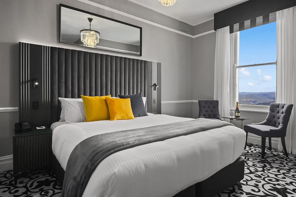 A modern hotel room with a large bed, a chair, a small table with a bottle, and a view of the outdoors through a window.