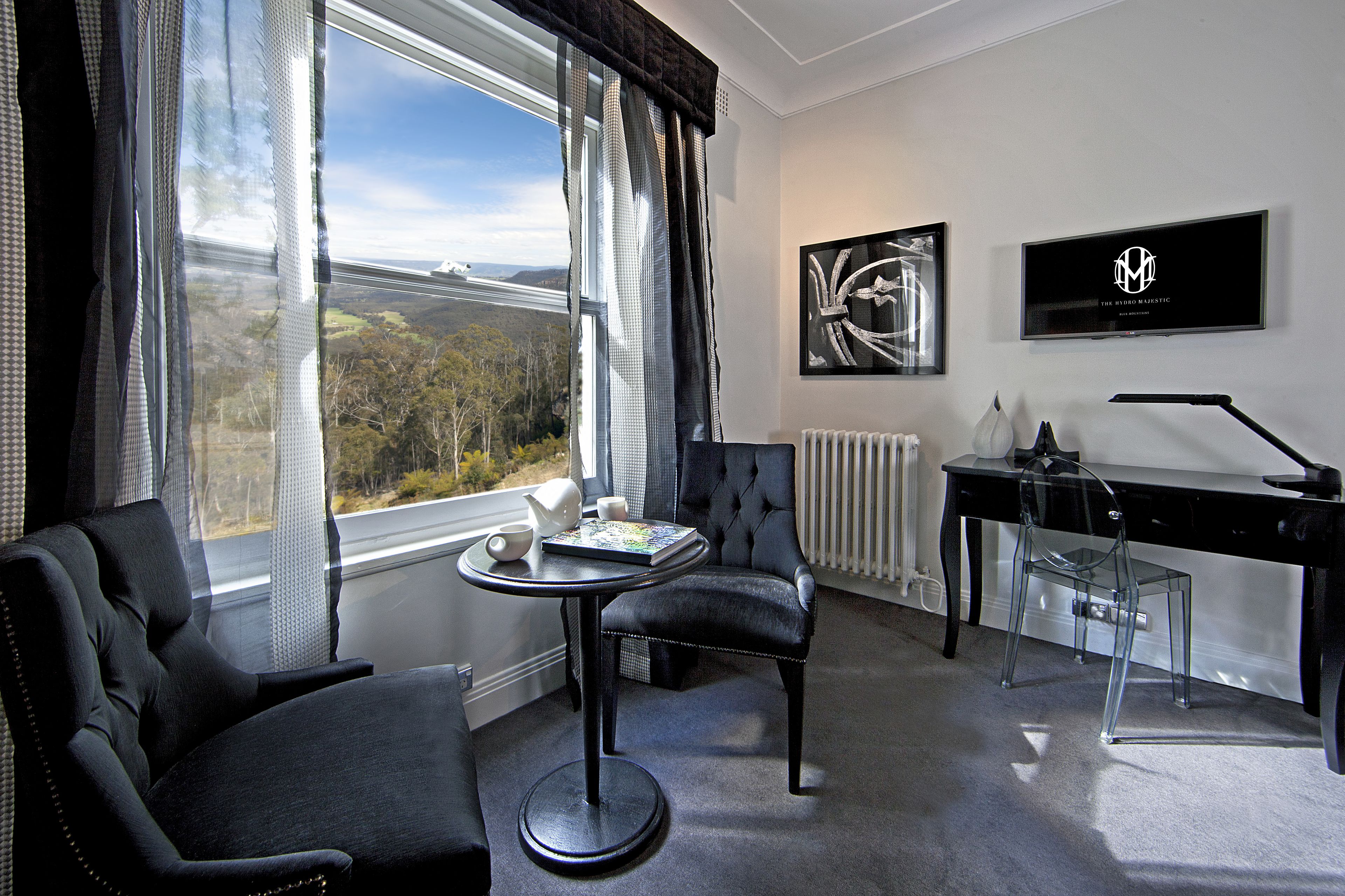 A modern room with a view of a bridge and trees, a black desk, a clear chair, and a round table with a book and teapot.