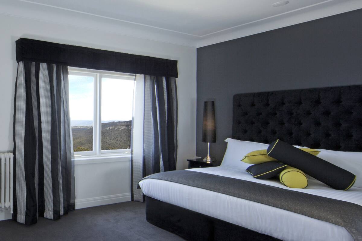 A bedroom with a large bed, a window with curtains, a nightstand with a lamp, and a view of the outdoors.