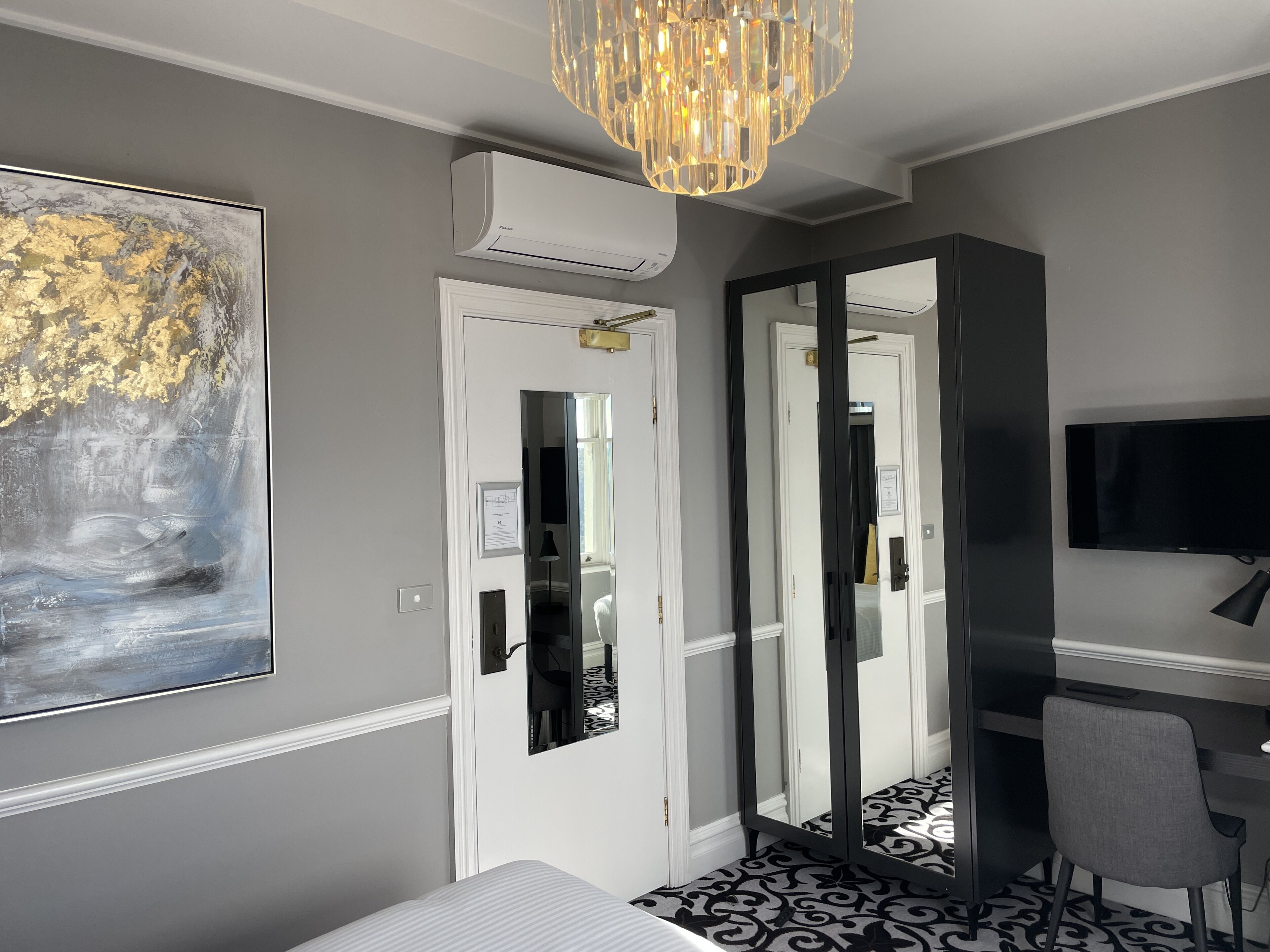 A modern hotel room with a large abstract painting, a chandelier, a flat-screen TV, and a desk with a chair.