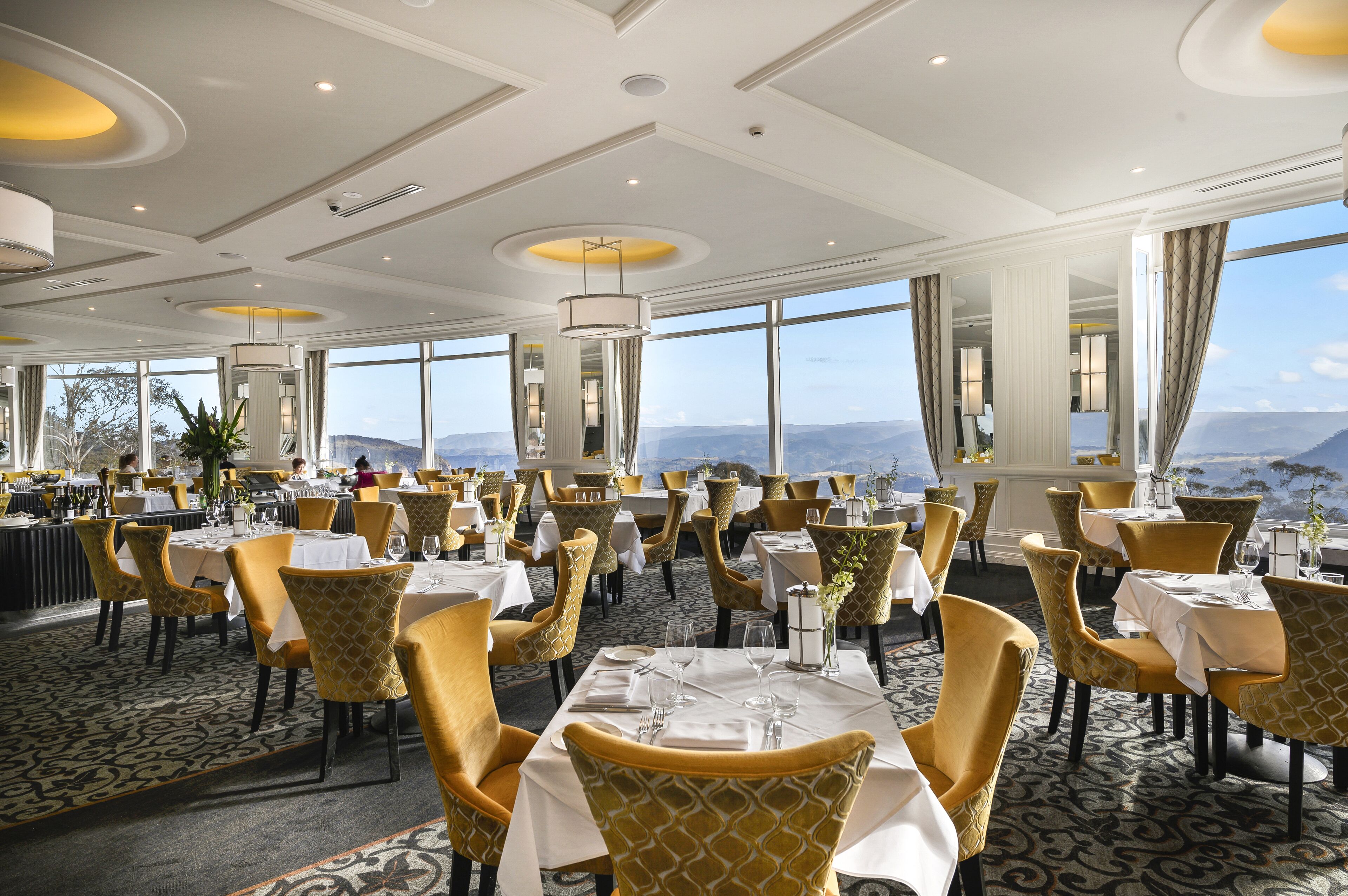 Un vaste espace dédié à la salle à manger, avec des tables et des chaises disposées pour accueillir les invités, de grandes fenêtres offrant une vue sur les montagnes et un design moderne au niveau du plafond.