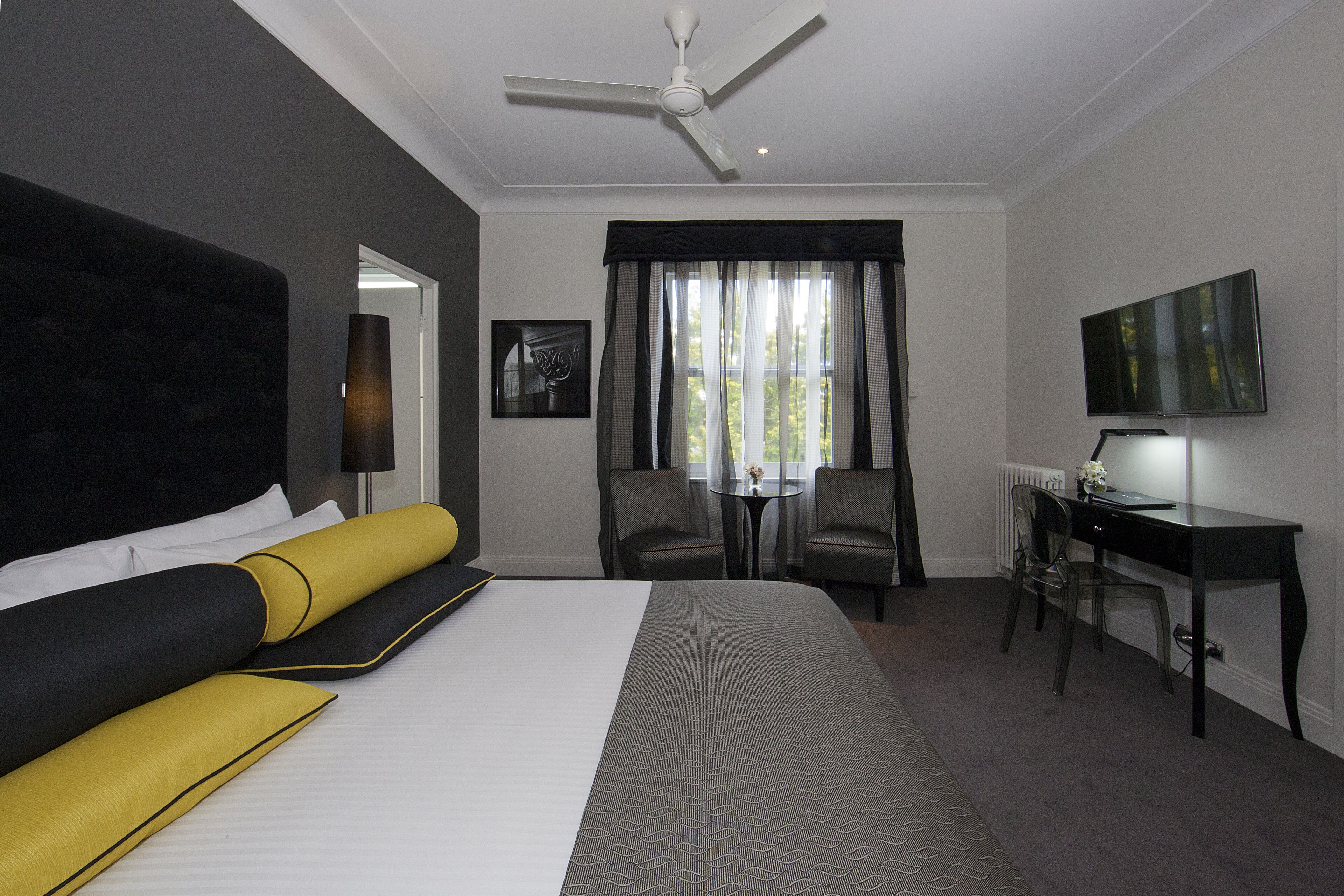 A hotel room with a large bed, a desk, two chairs, a ceiling fan, and a TV on the wall.