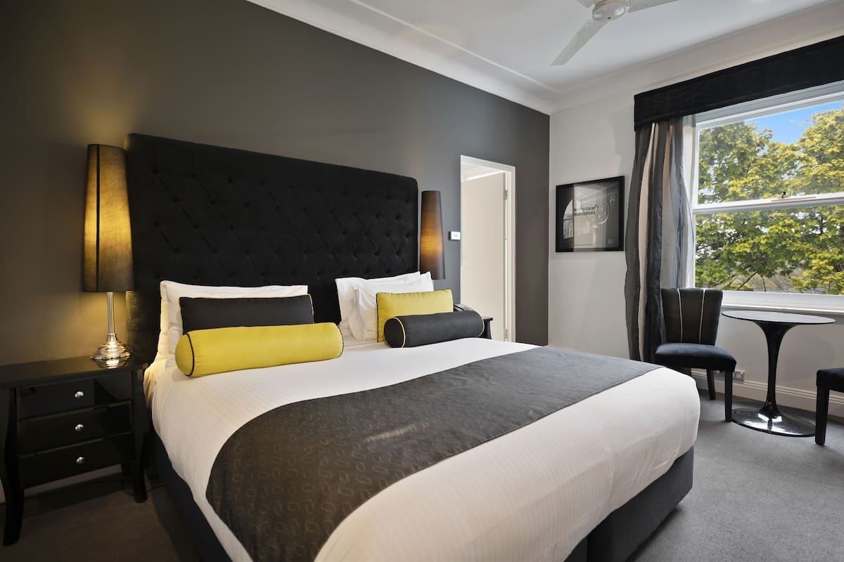 A hotel room with a large bed, a bedside table, a chair, a small table, and a window with a view of trees.