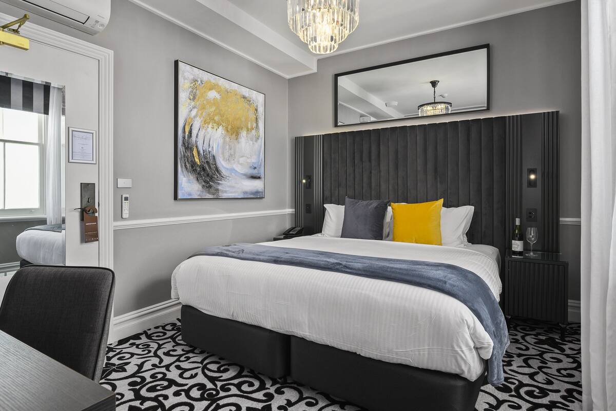 A modern hotel room with a large bed, a bedside table with a wine glass, a mirror, and a painting on the wall.