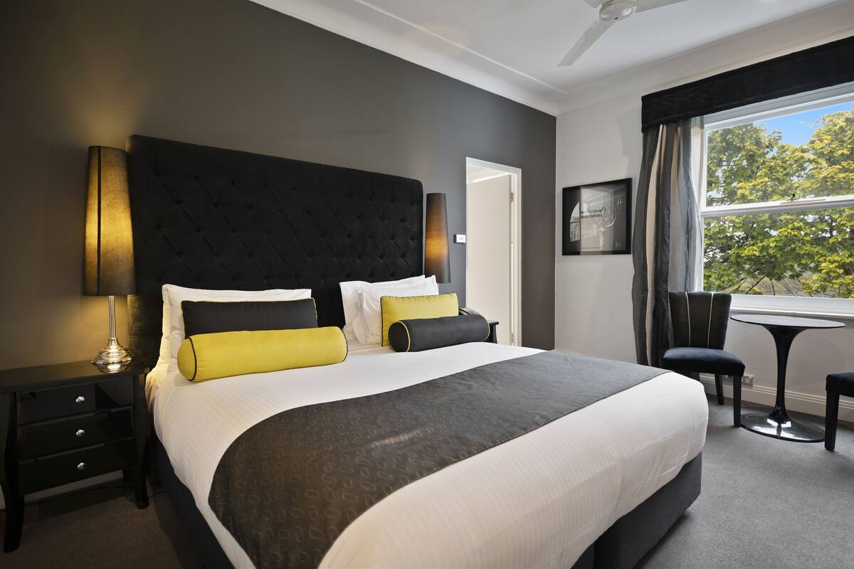A hotel room with a large bed, a bedside table, a chair, a small table, and a window with a view of trees.