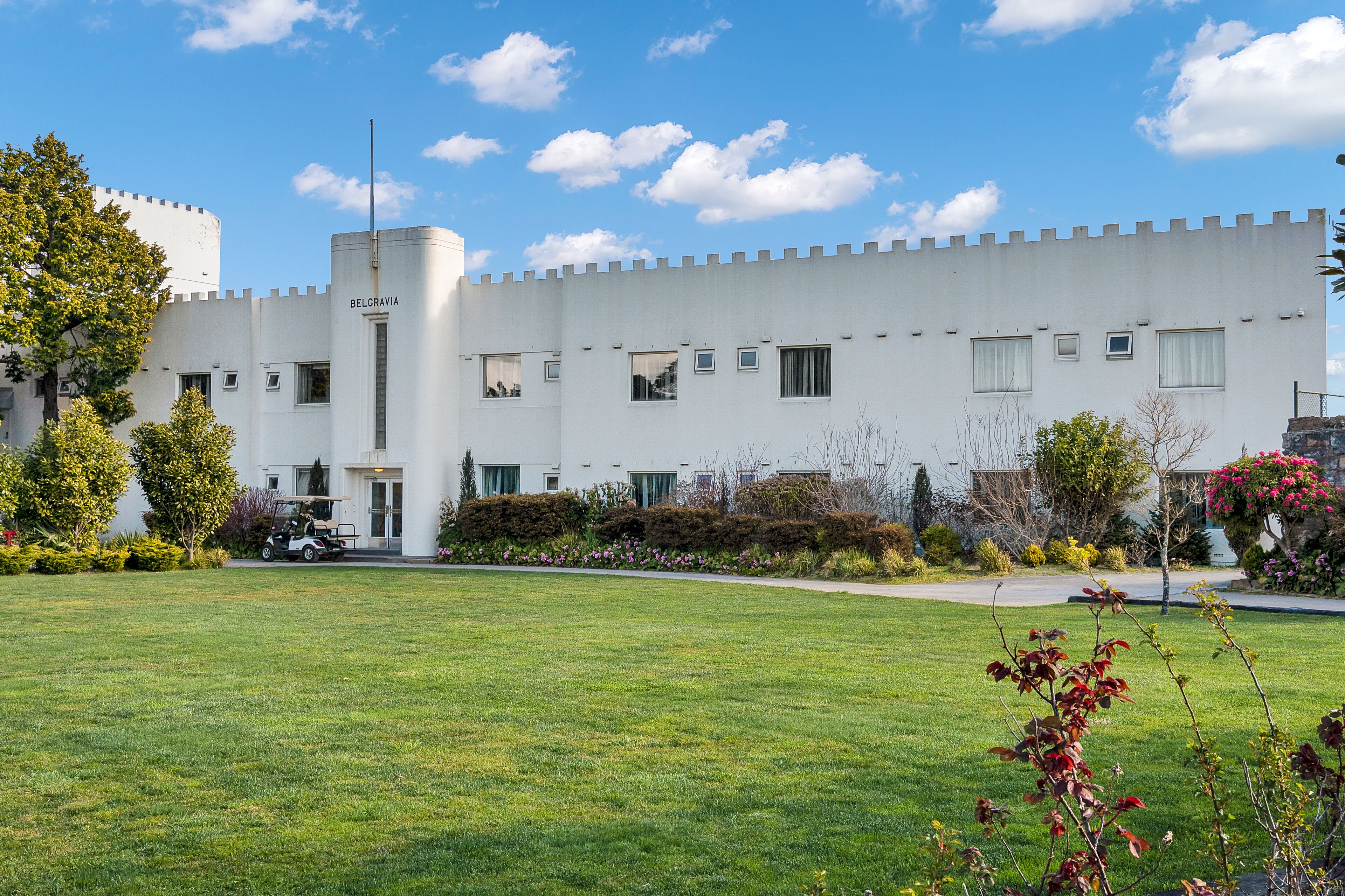 A white building with a turret and multiple windows, surrounded by a well-maintained lawn and various plants.