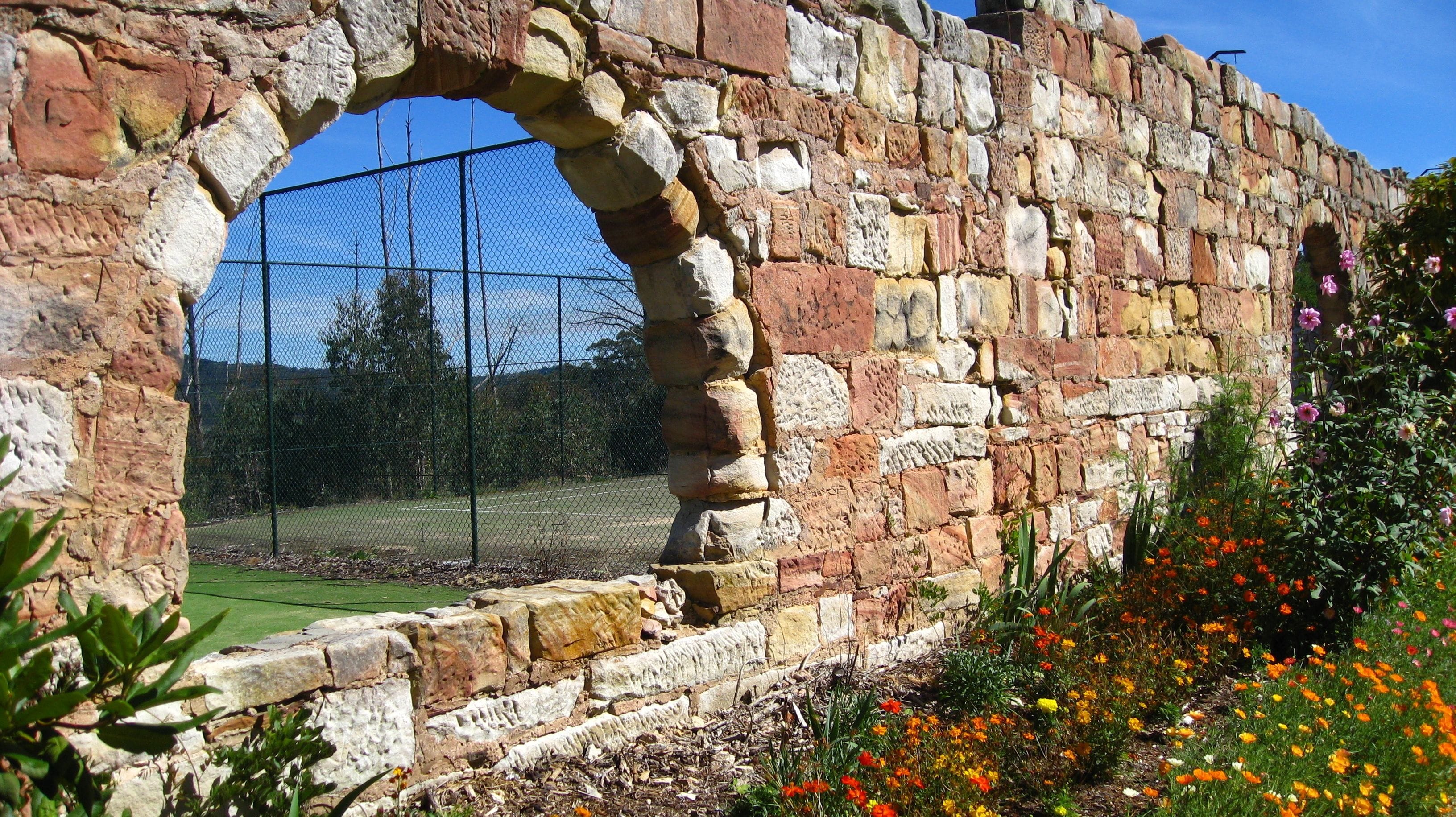 An ancient stone wall with a large arched opening, a chain-link fence, and a garden with colorful flowers.