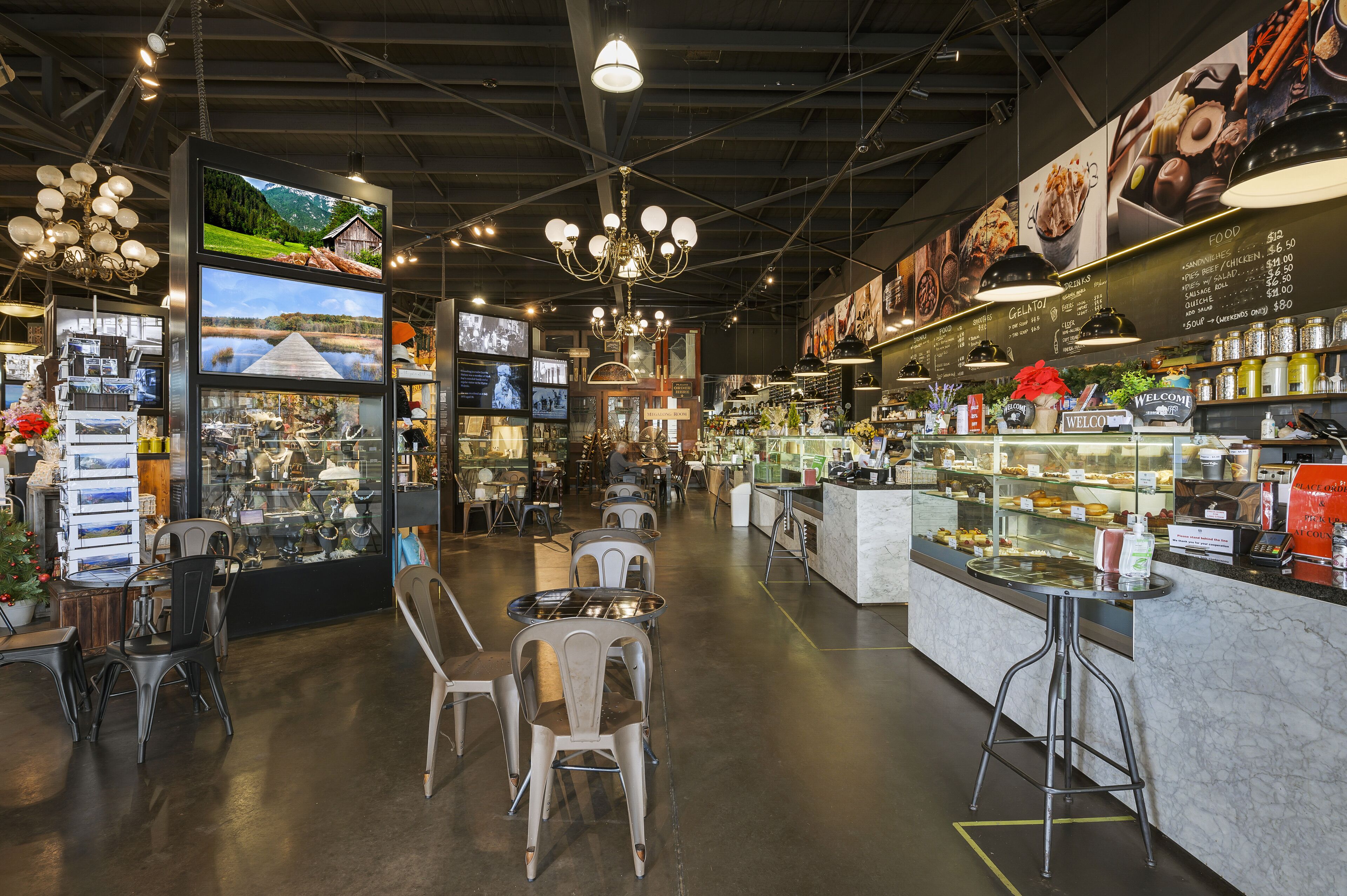 Een modern café met een centrale zithoek, een balie met een menukaart, en diverse etalages met eten.