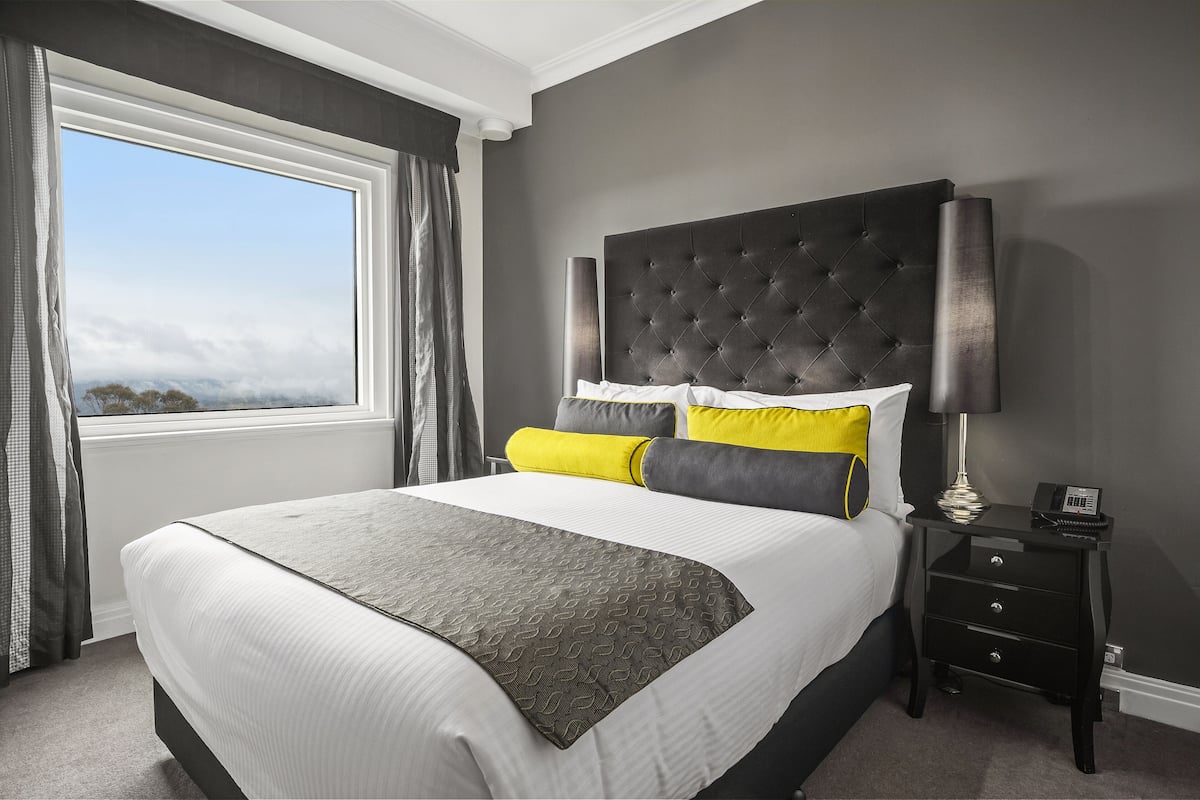 A neatly made bed with a grey and white duvet, a black headboard, and yellow and grey pillows. There is a bedside table with a lamp and a phone, and a window with curtains.
