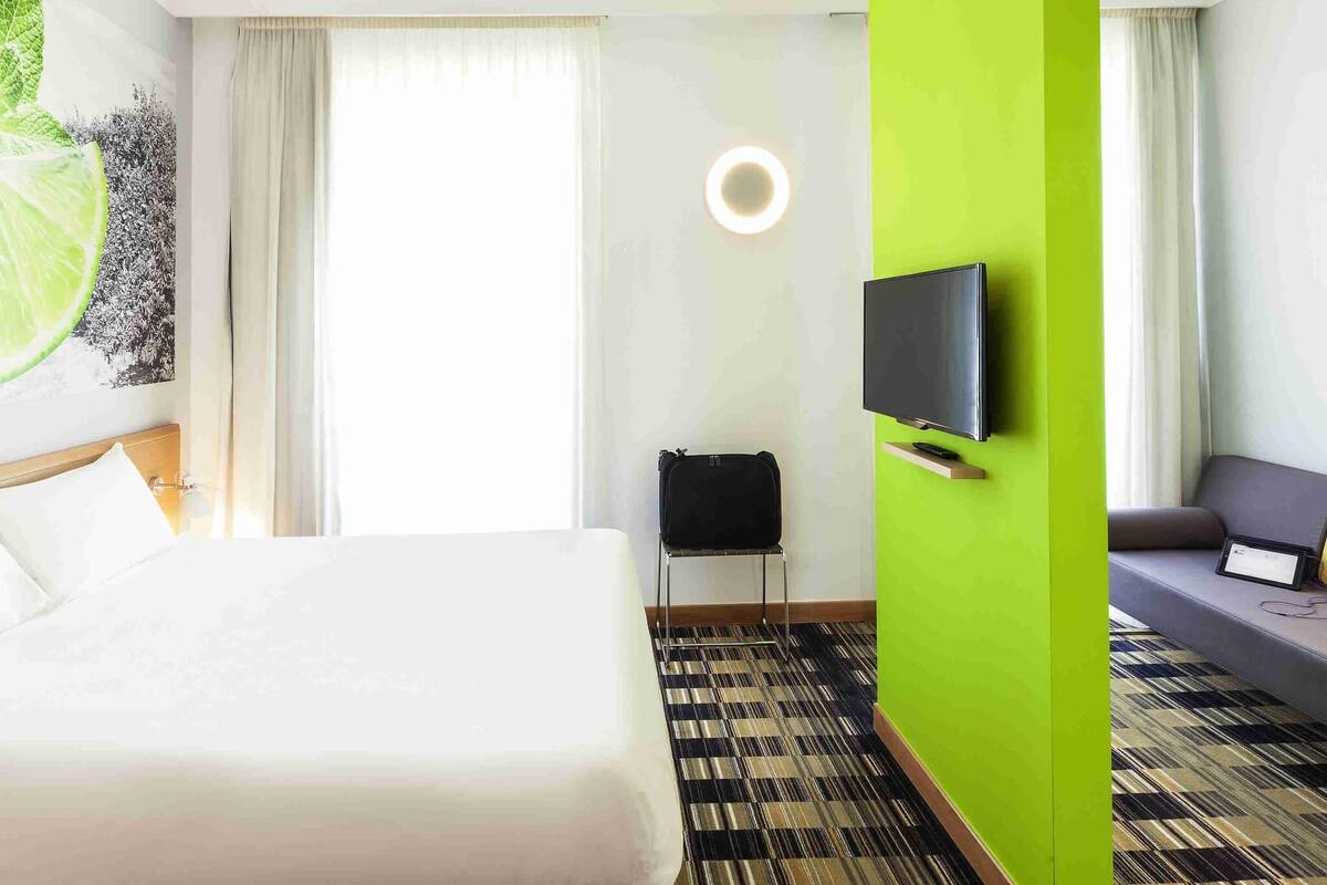 Ein Hotelzimmer mit einem Bett, einem an der Wand montierten Fernseher, einer Couch und einem gemusterten Teppich.