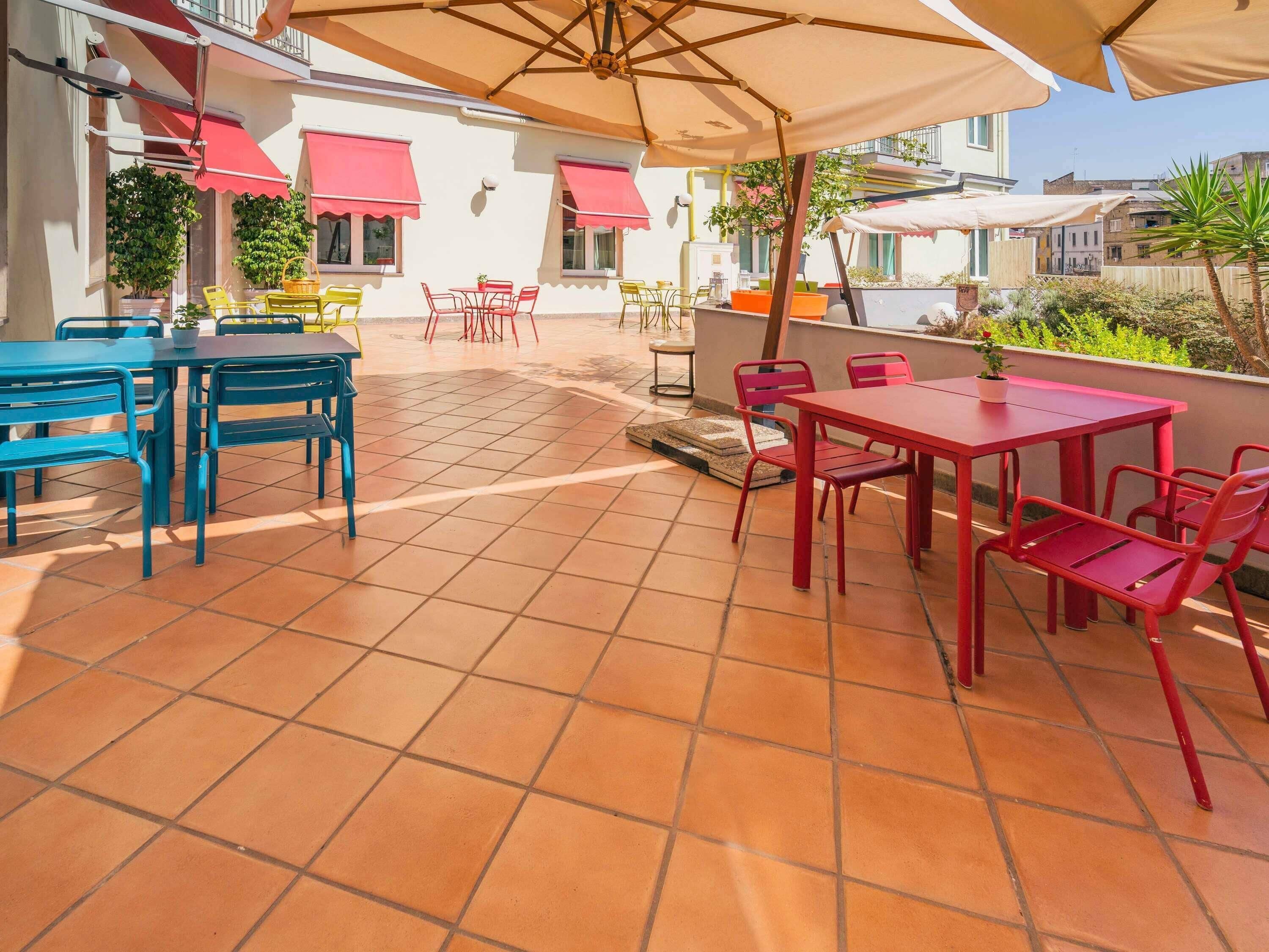 Un patio al aire libre con suelo de baldosas, mobiliario exterior rojo y azul, y sombrillas que dan sombra.