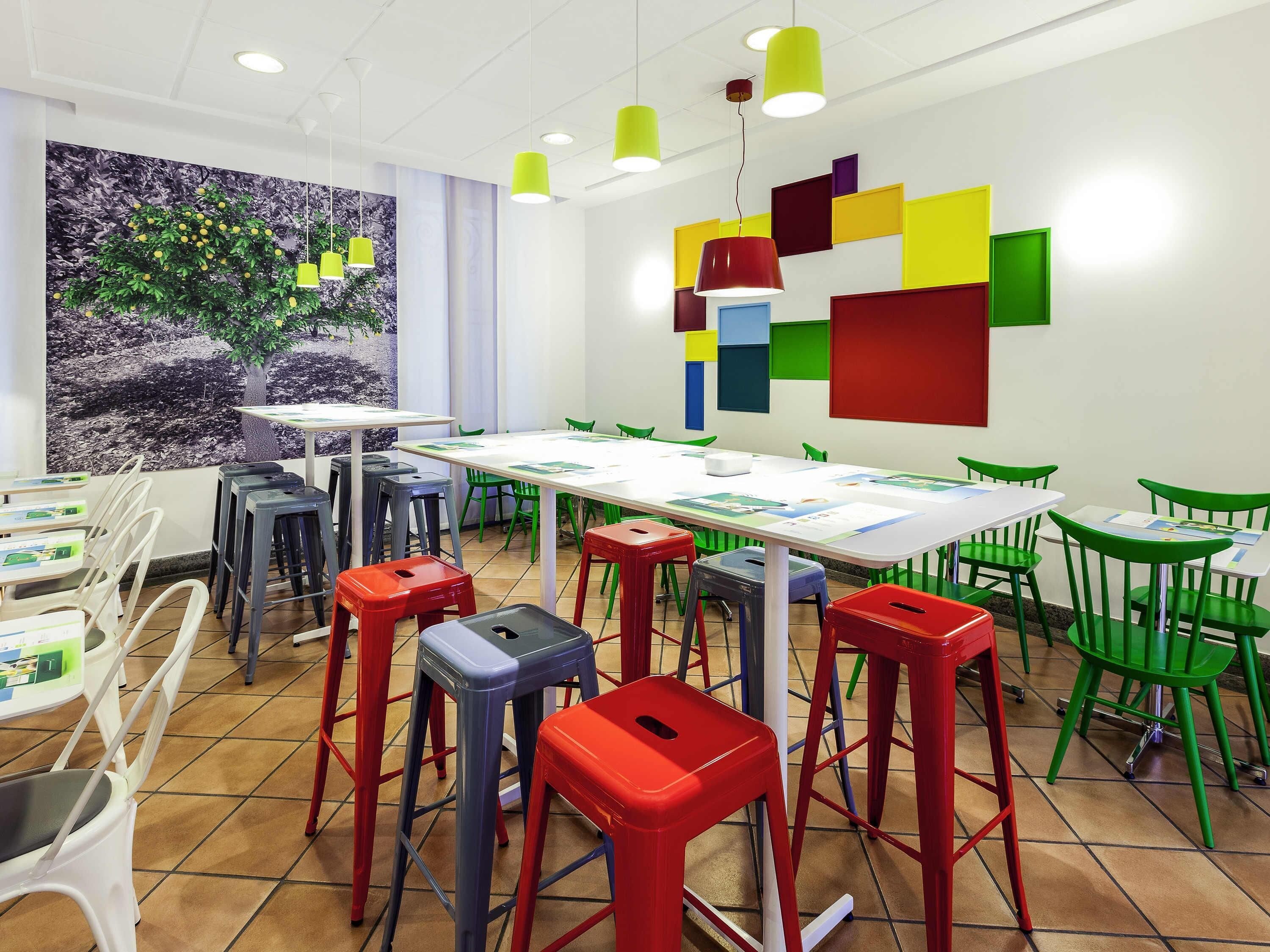 A brightly lit room with a long table, colorful stools, and a mural on the wall.