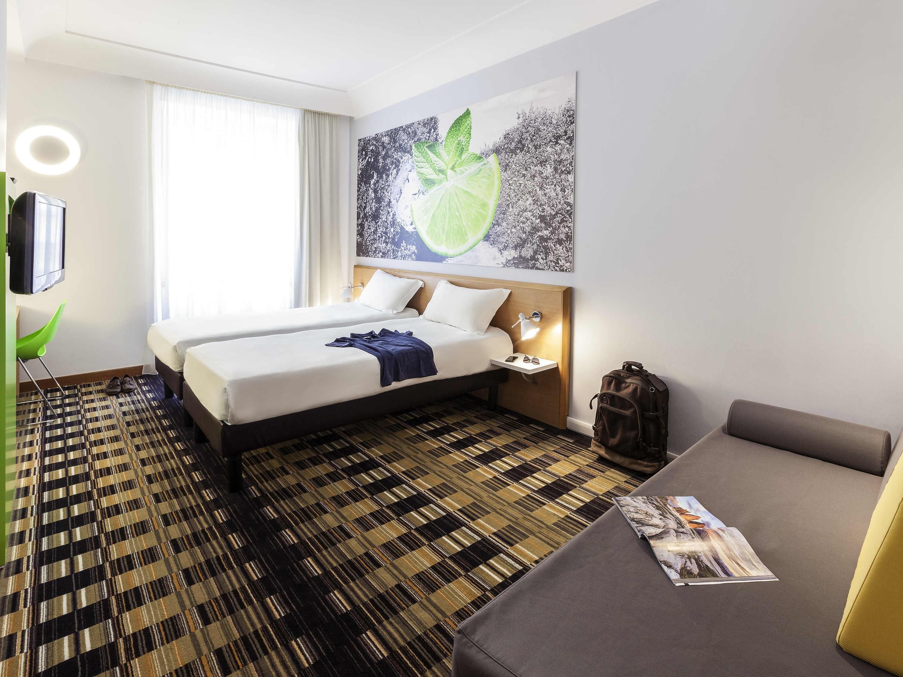 Ein Hotelzimmer mit einem Bett, einem Nachttisch, einer Lampe, einem Fernseher, einem grünen Sessel, einem Sofa und einem gemusterten Teppich.