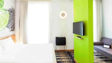 Ein Hotelzimmer mit Bett, Wandfernseher, Sofa, kleinem Tisch und Koffer auf dem Boden.
