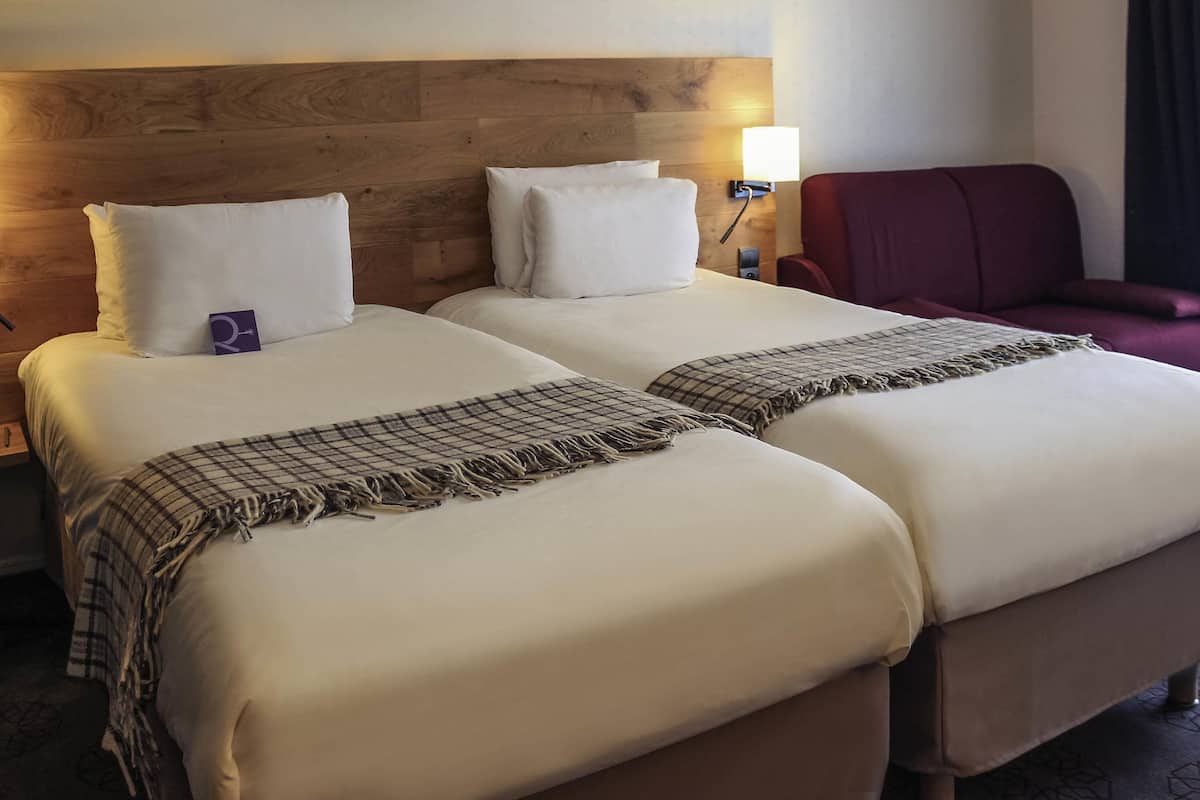 Una habitación de hotel con dos camas, un cabecero de madera, una silla roja y una lámpara fijada a la pared.