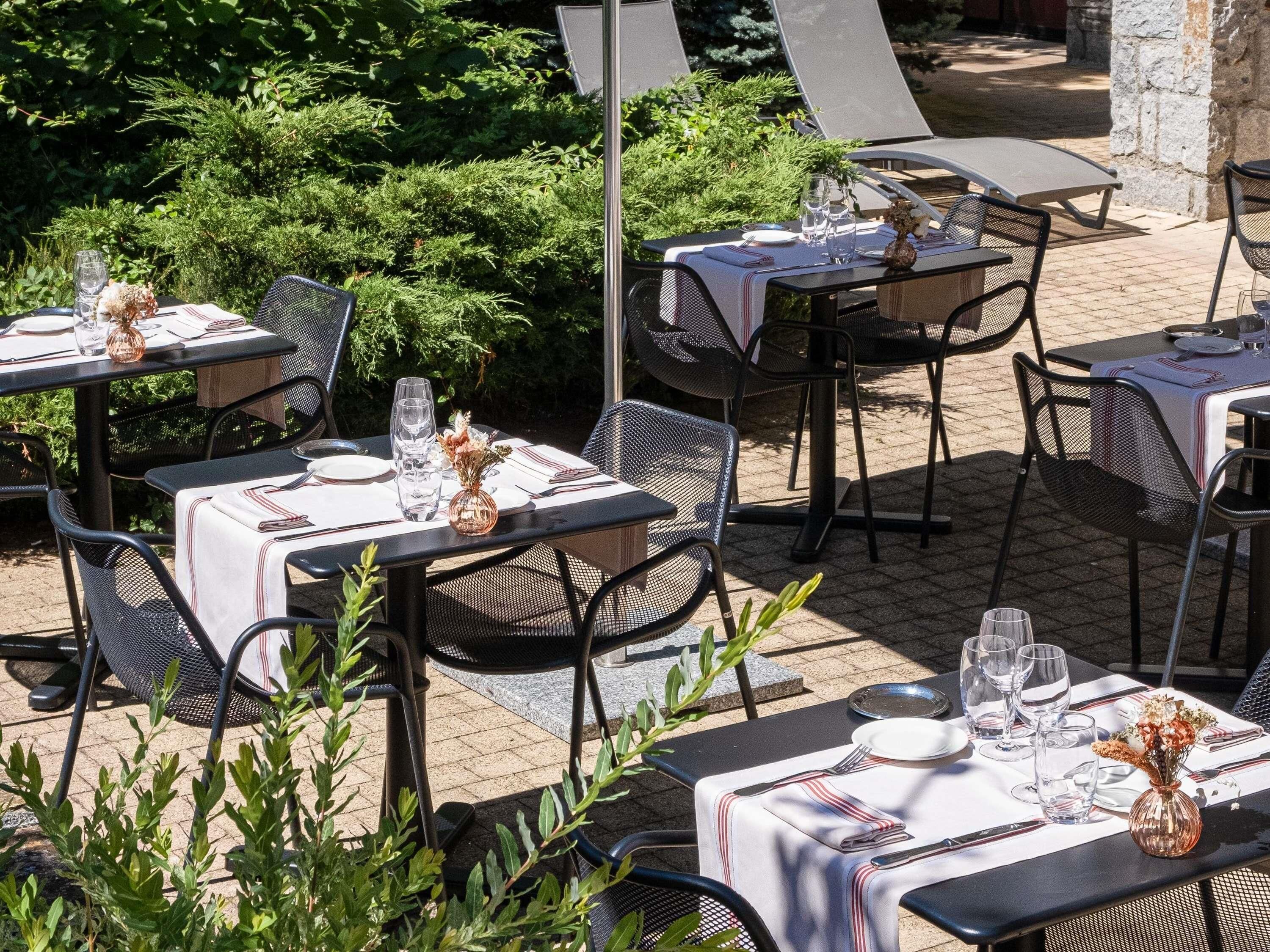 Mesas de comedor al aire libre con sillas, dispuestas con platos, vasos y cubiertos, rodeadas de follaje.