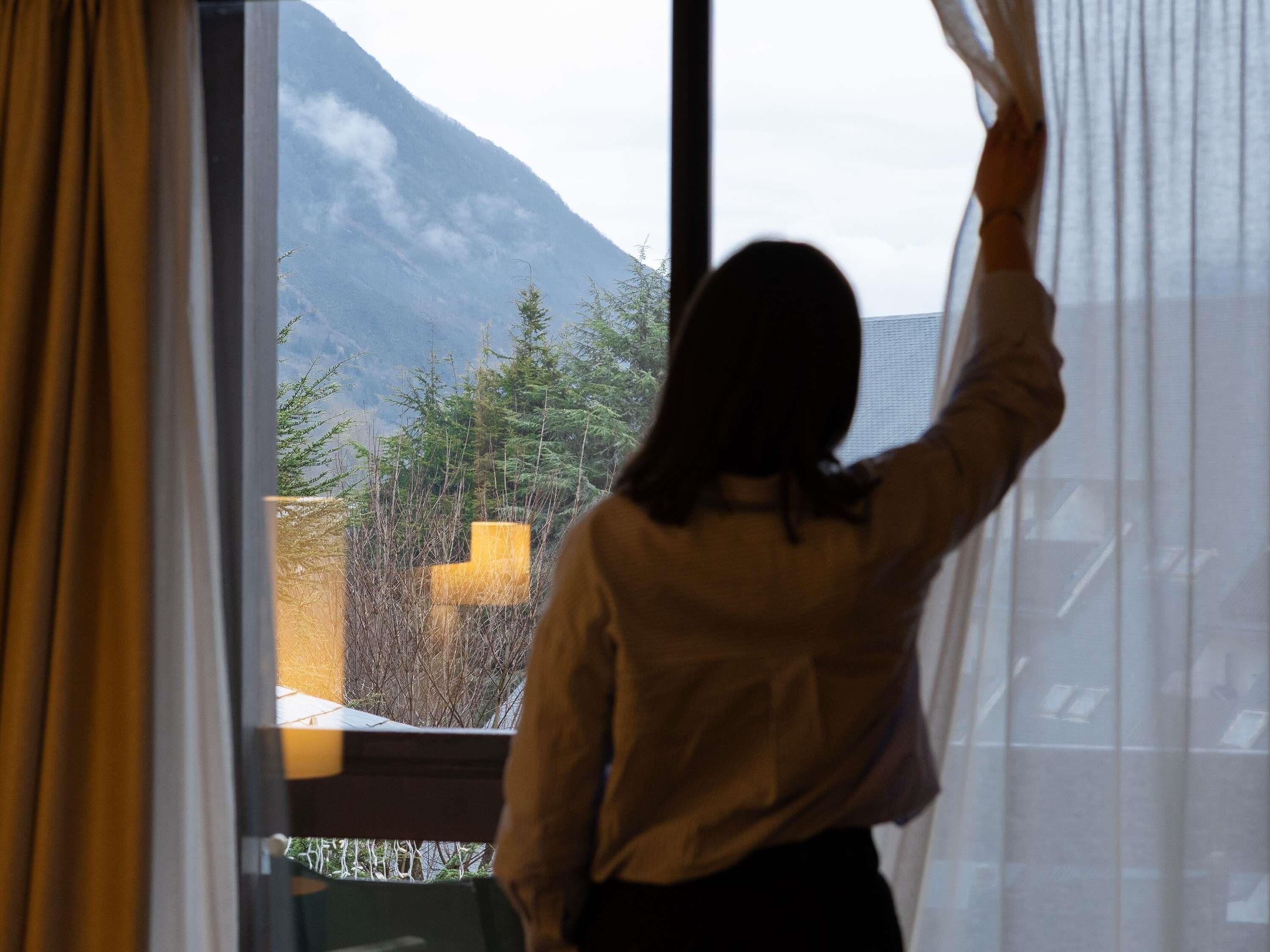 Una persona parada junto a una ventana con cortinas, mirando hacia una montaña y árboles.