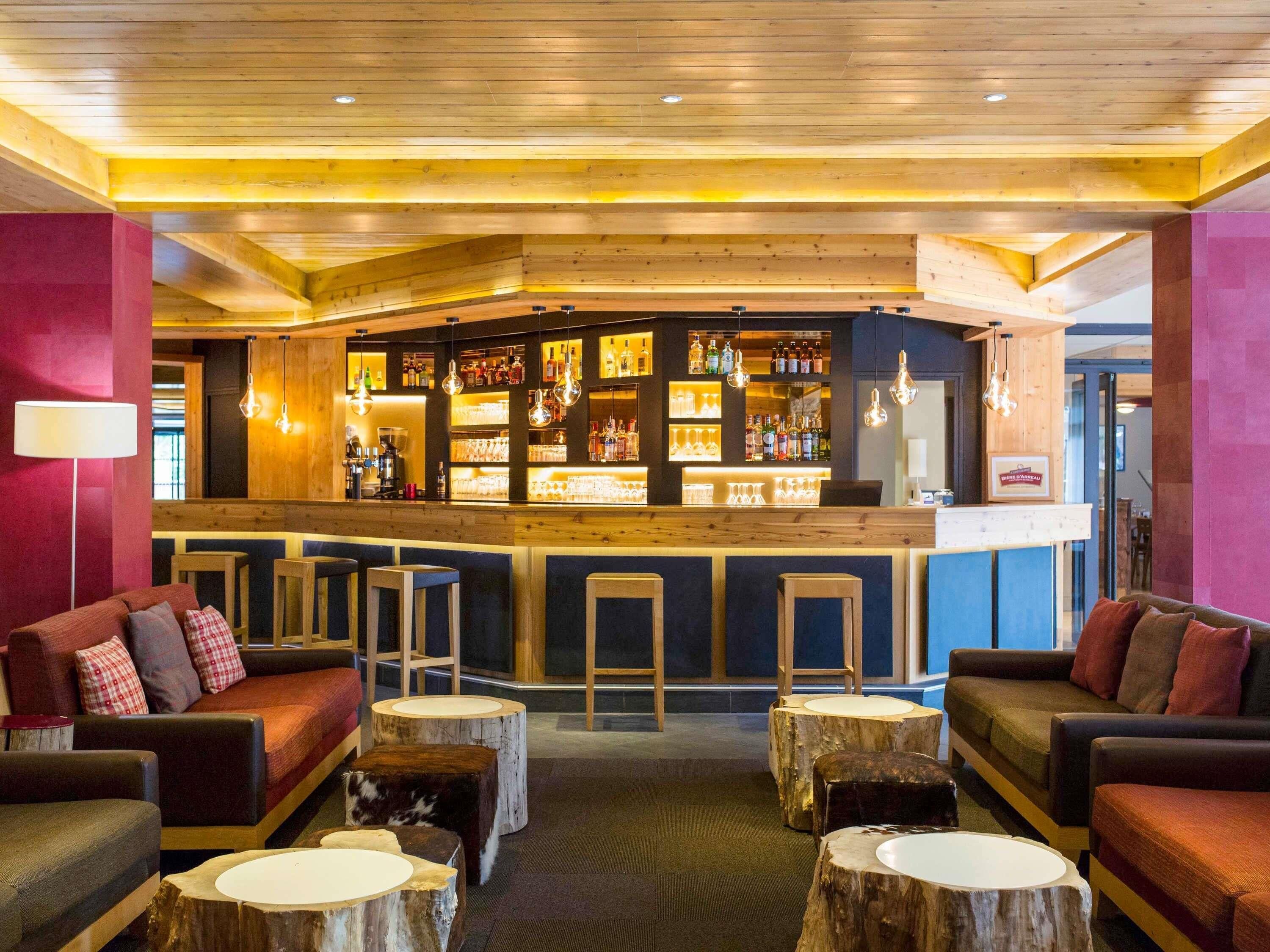 A bar area with wooden bar stools, a counter with bottles, and seating with round tables.